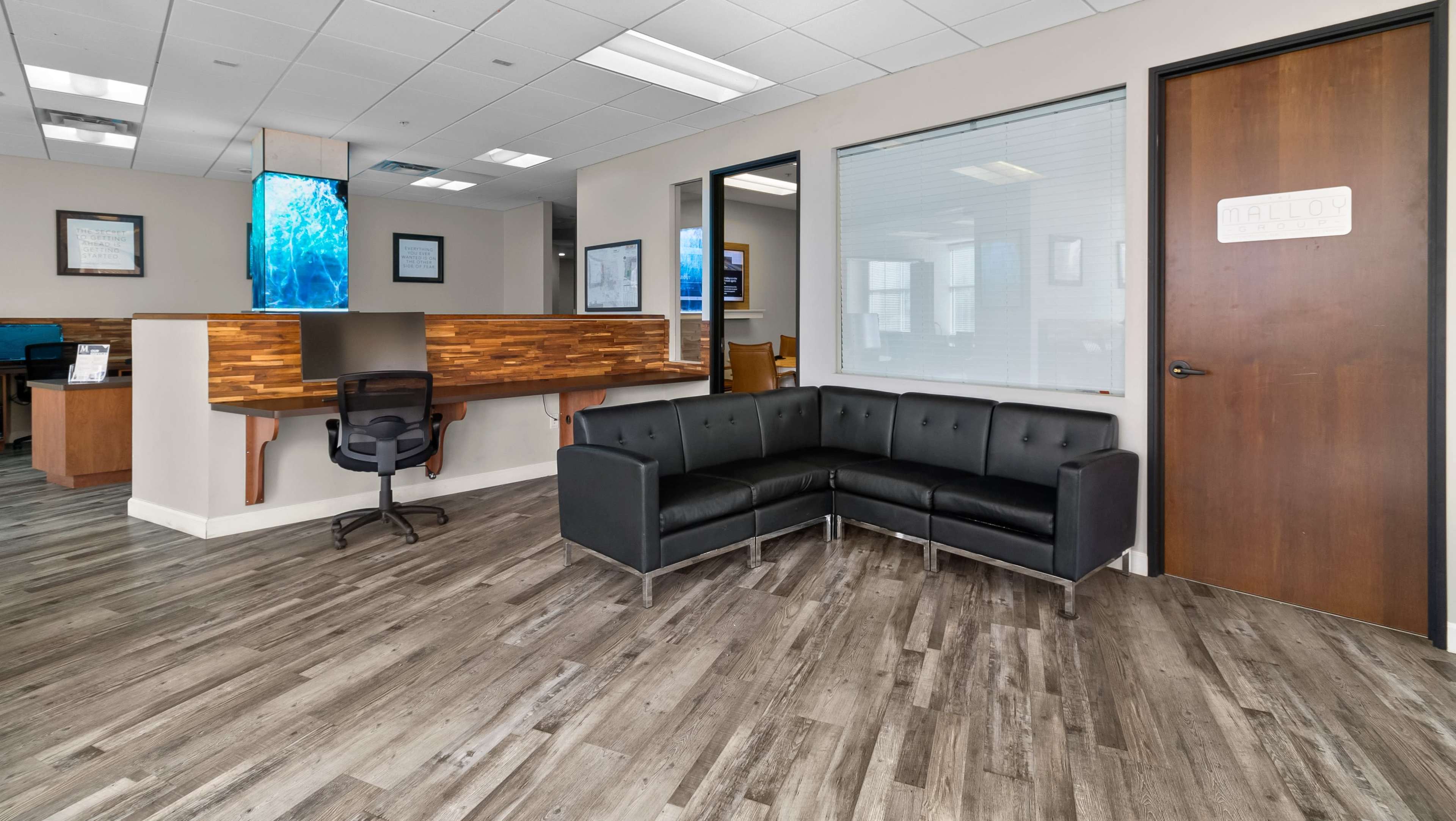 The image shows a modern office reception area with a black sectional sofa, wooden counter, and large windows allowing natural light.