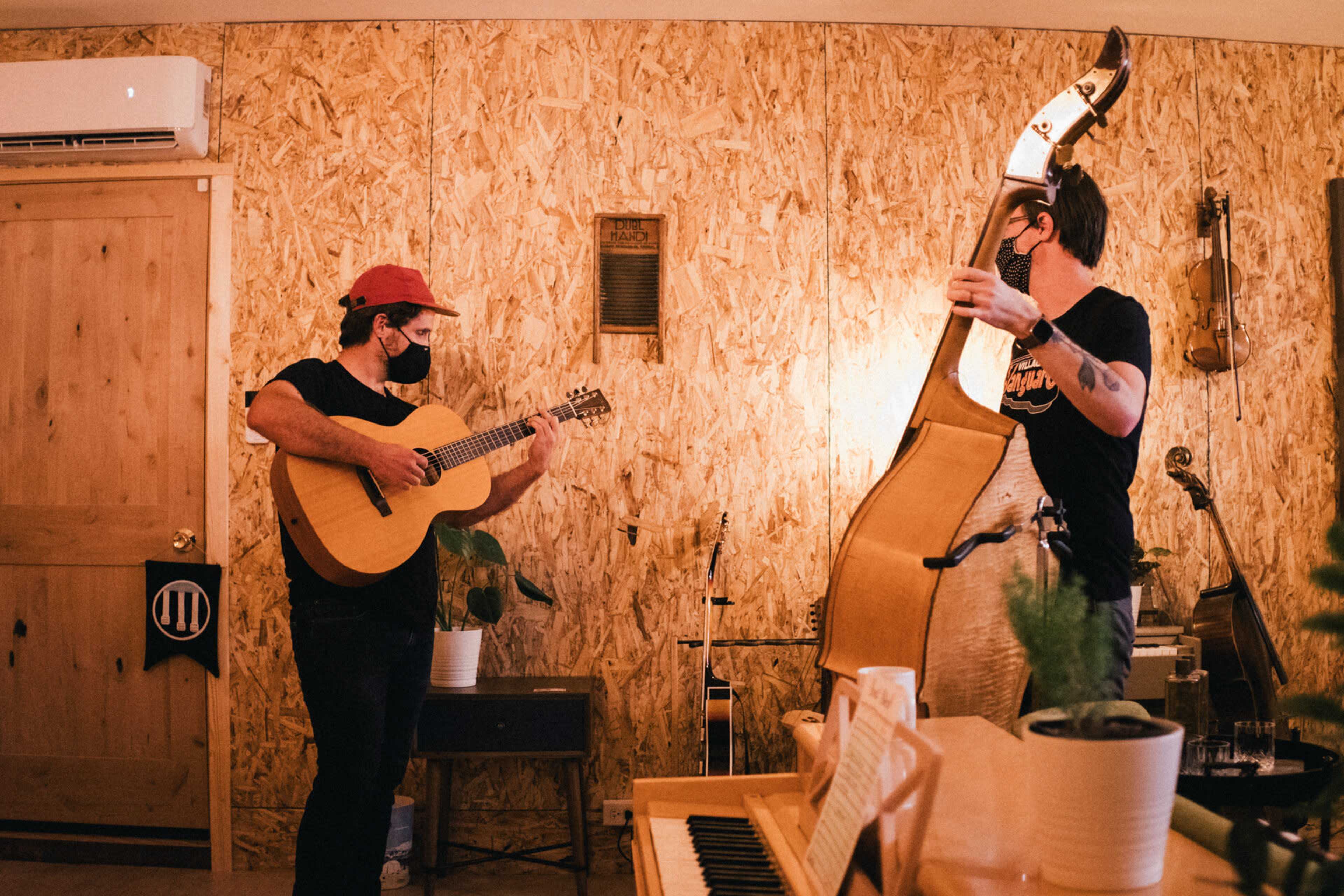 Two musicians play instruments—a guitarist holding an acoustic guitar and a bassist—with wooden walls and various musical instruments in the background.