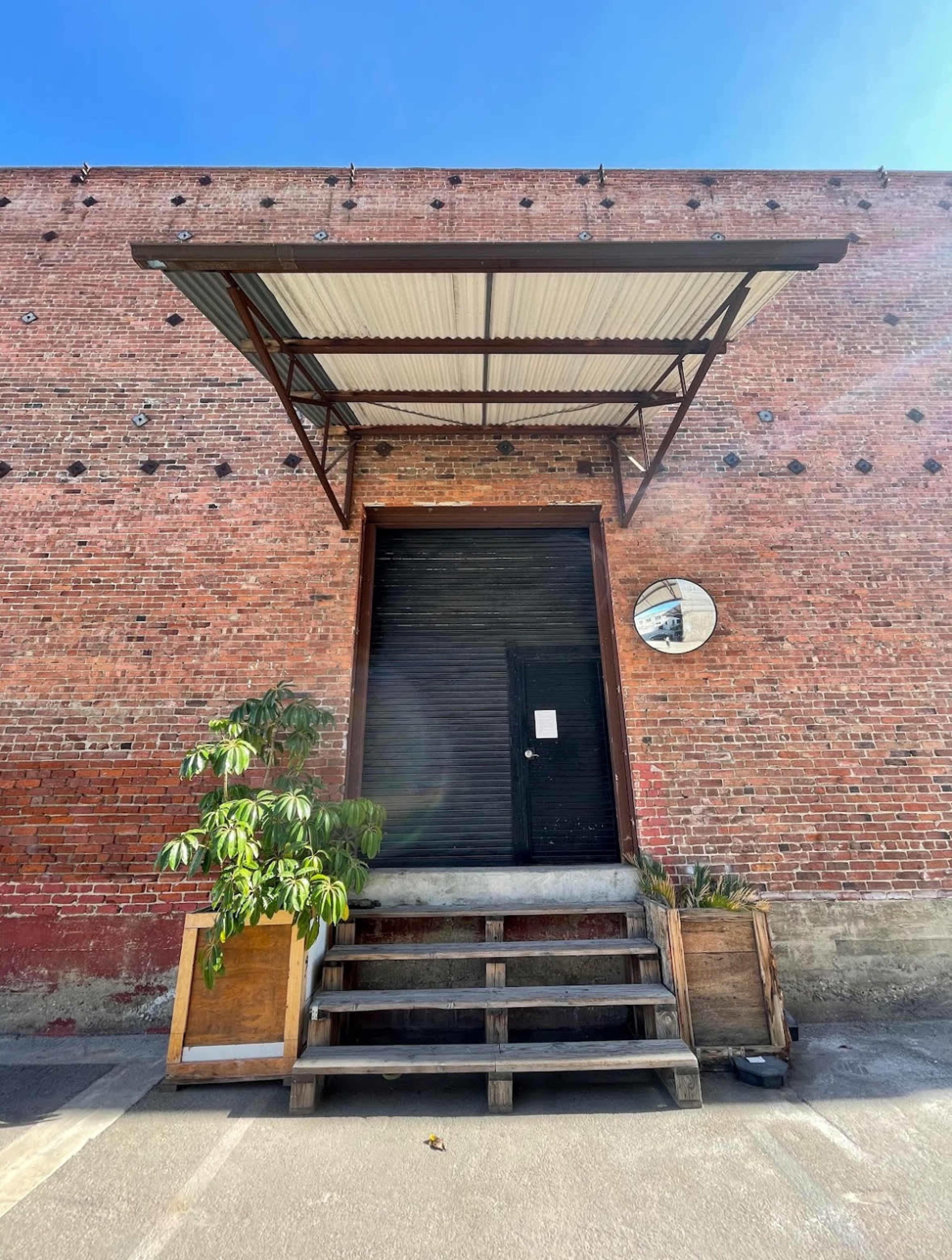 A dark door with a metal awning and a round mirror is set against a brick wall, surrounded by wooden steps and potted plants.