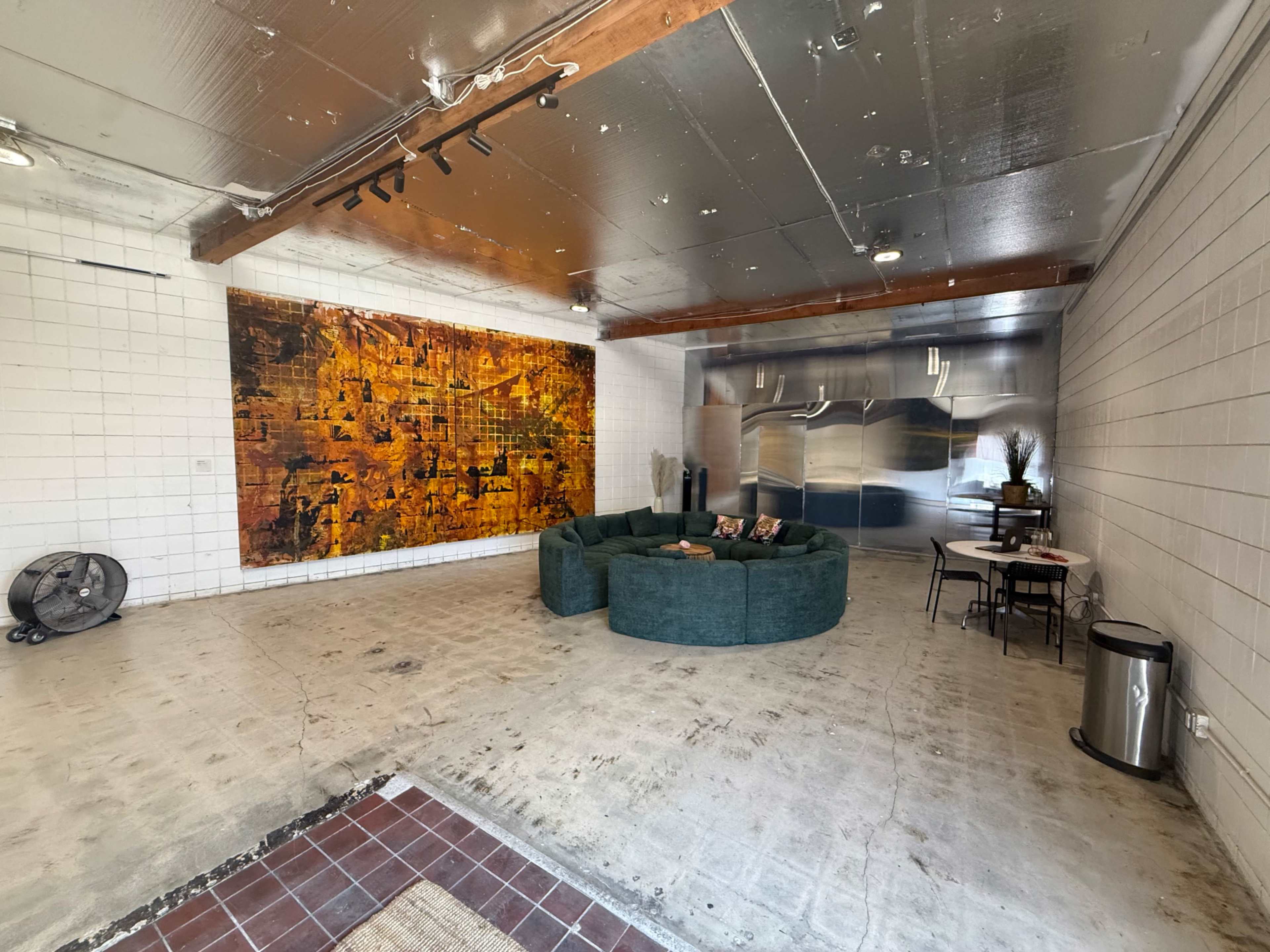 The image shows a spacious room featuring a large circular green sofa, a wall with an abstract artwork, a kitchen area with stainless steel appliances, and a small dining table.