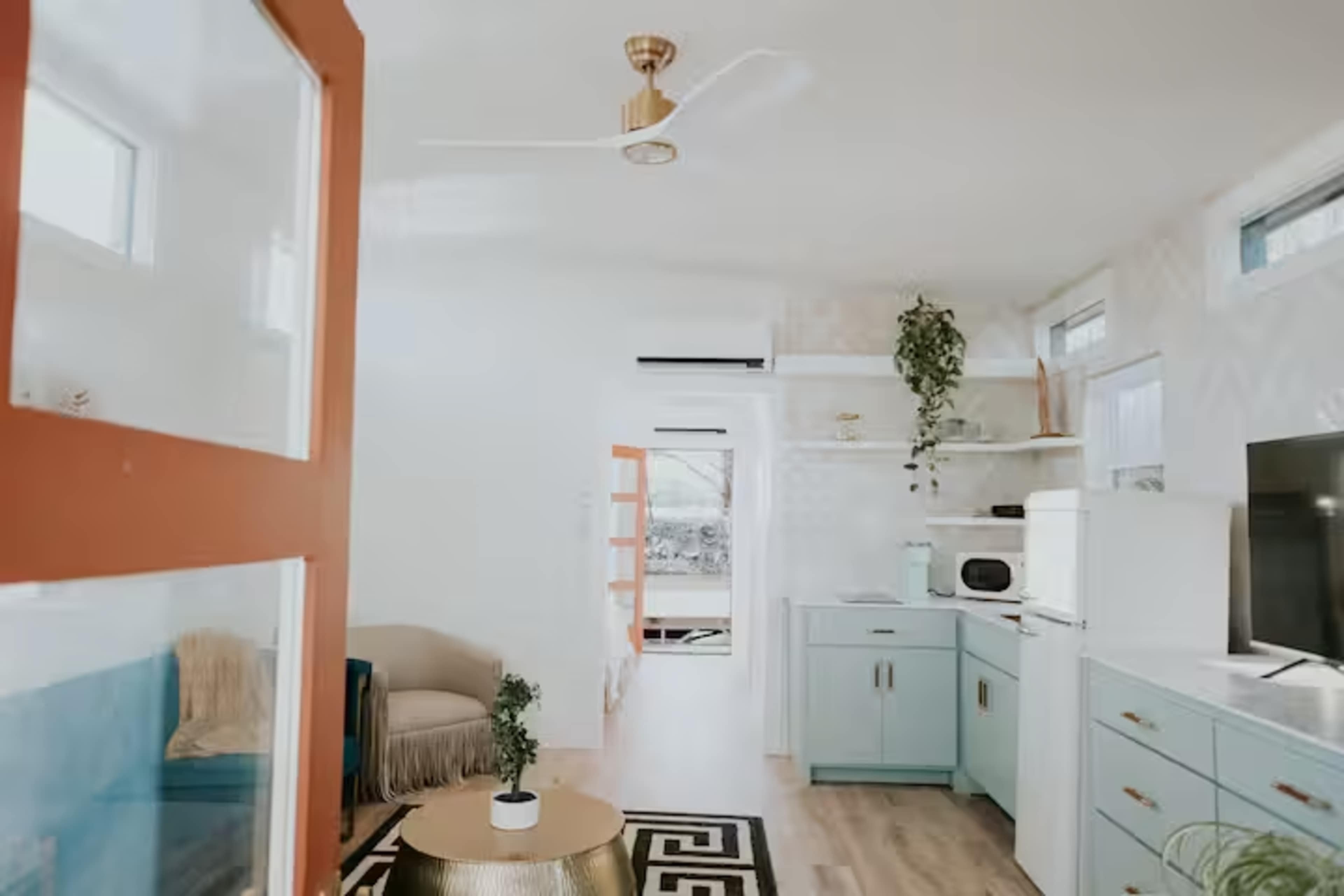 The image shows a modern, open-plan living area featuring a kitchen with light blue cabinetry, a dining space, and a cozy seating area with a patterned rug.