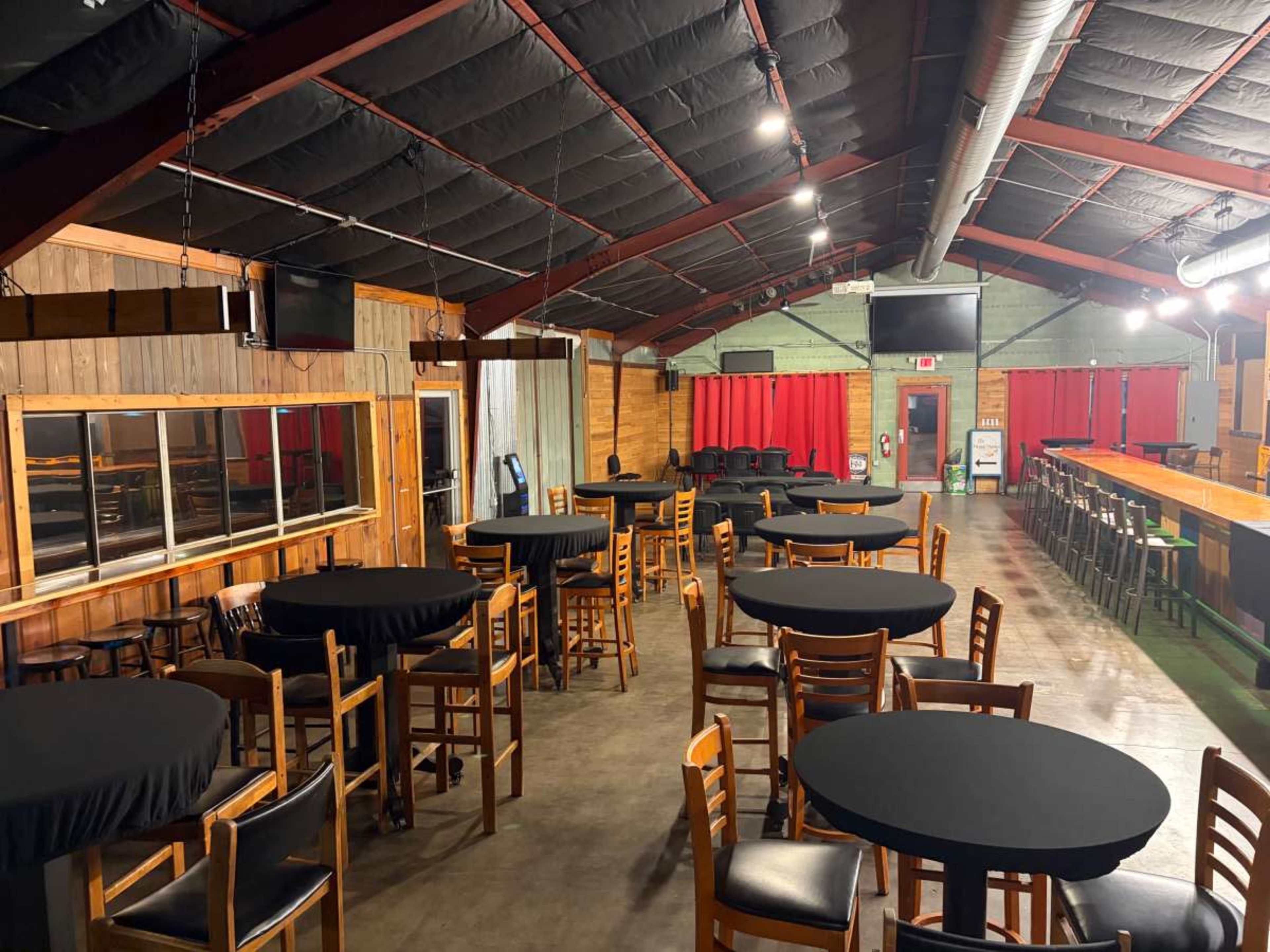 The image shows a spacious indoor venue with wooden walls, several round tables covered in black cloth, and a bar area at one end, illuminated by overhead lights.