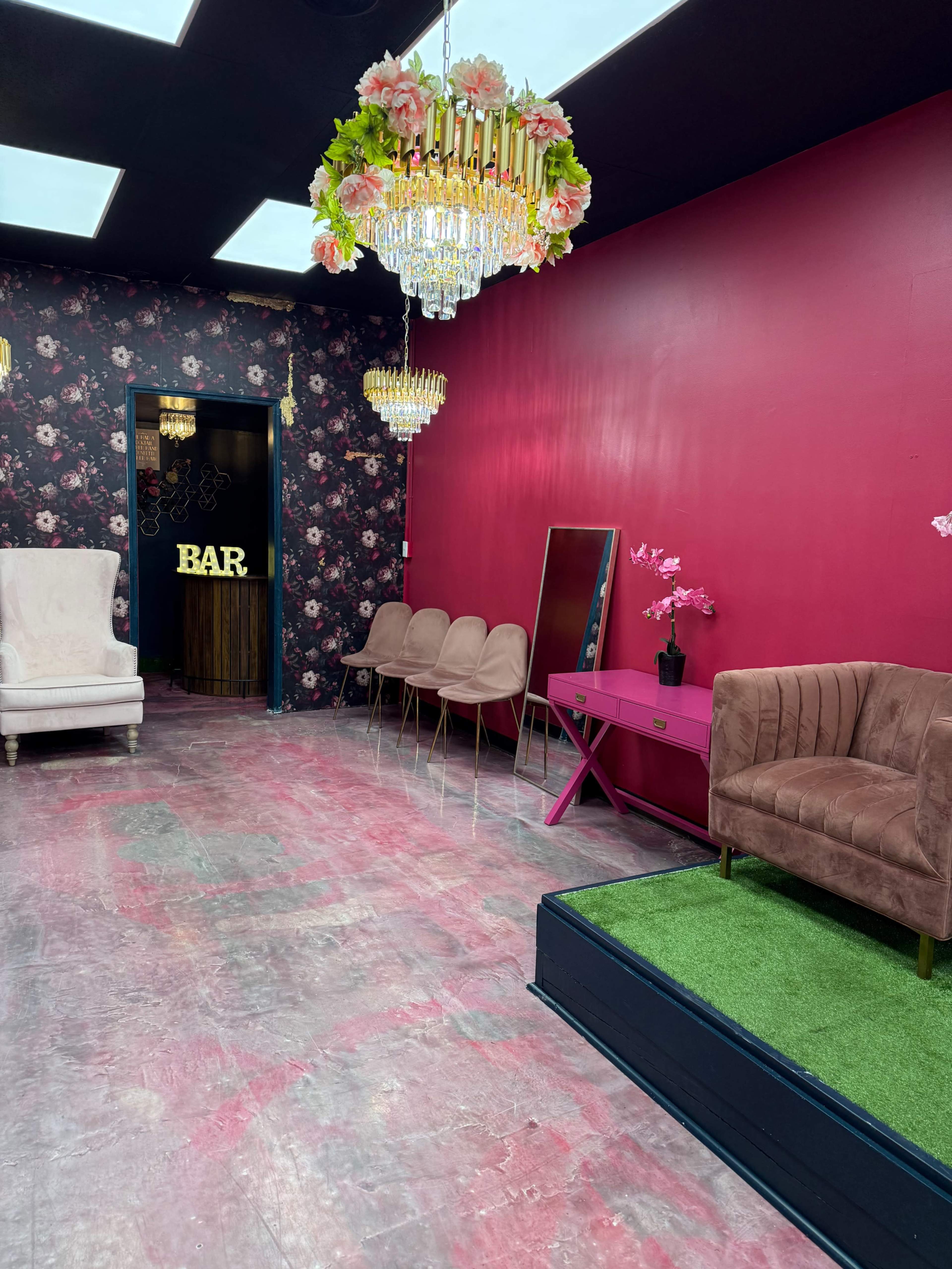 The room features pink walls adorned with floral wallpaper, a bar sign in the back, elegant chandeliers, and a mix of seating arrangements.