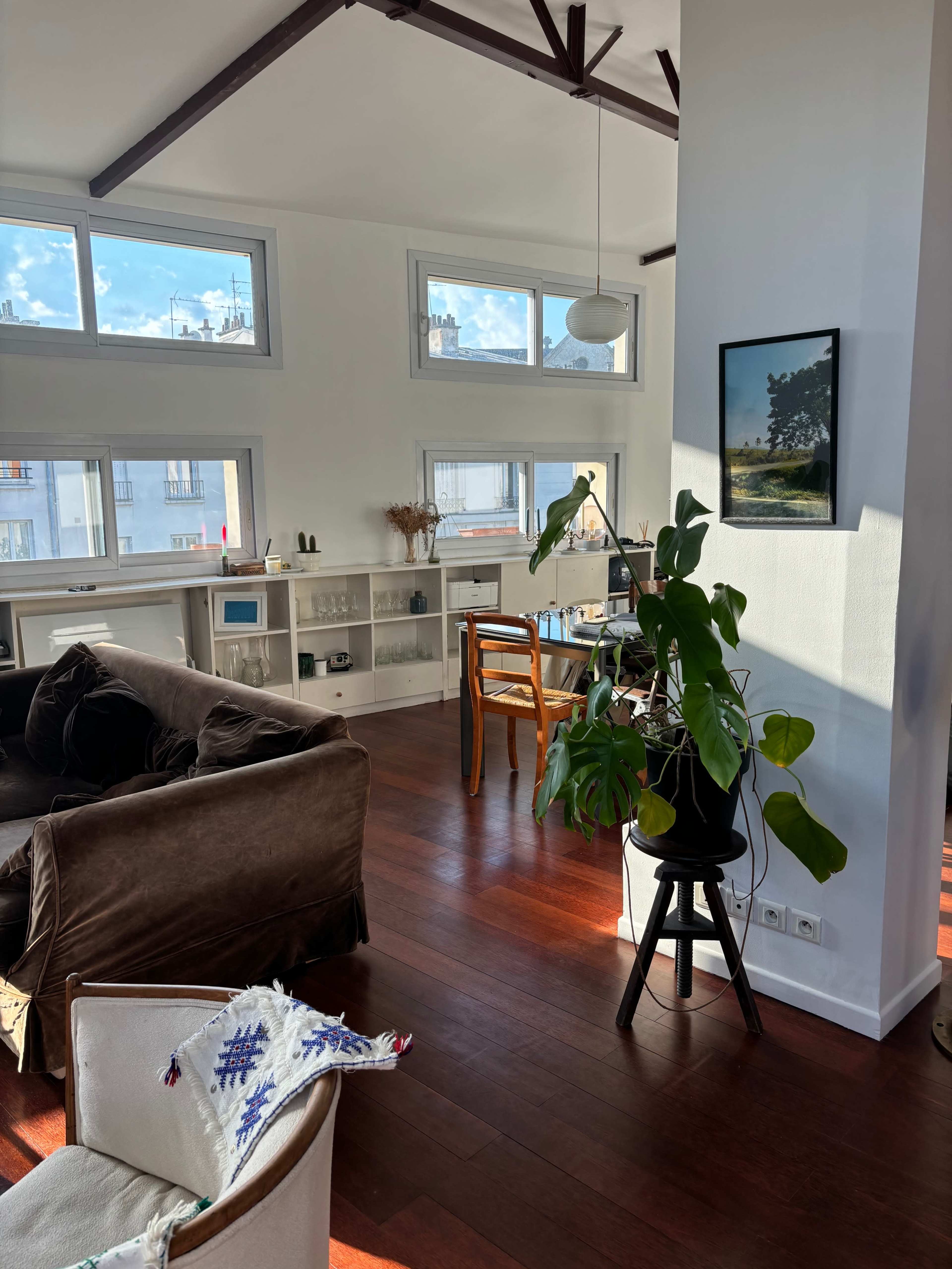 The image shows a bright, modern living space with hardwood floors, large windows, a potted plant, and a mix of furniture including a sofa and dining chairs.