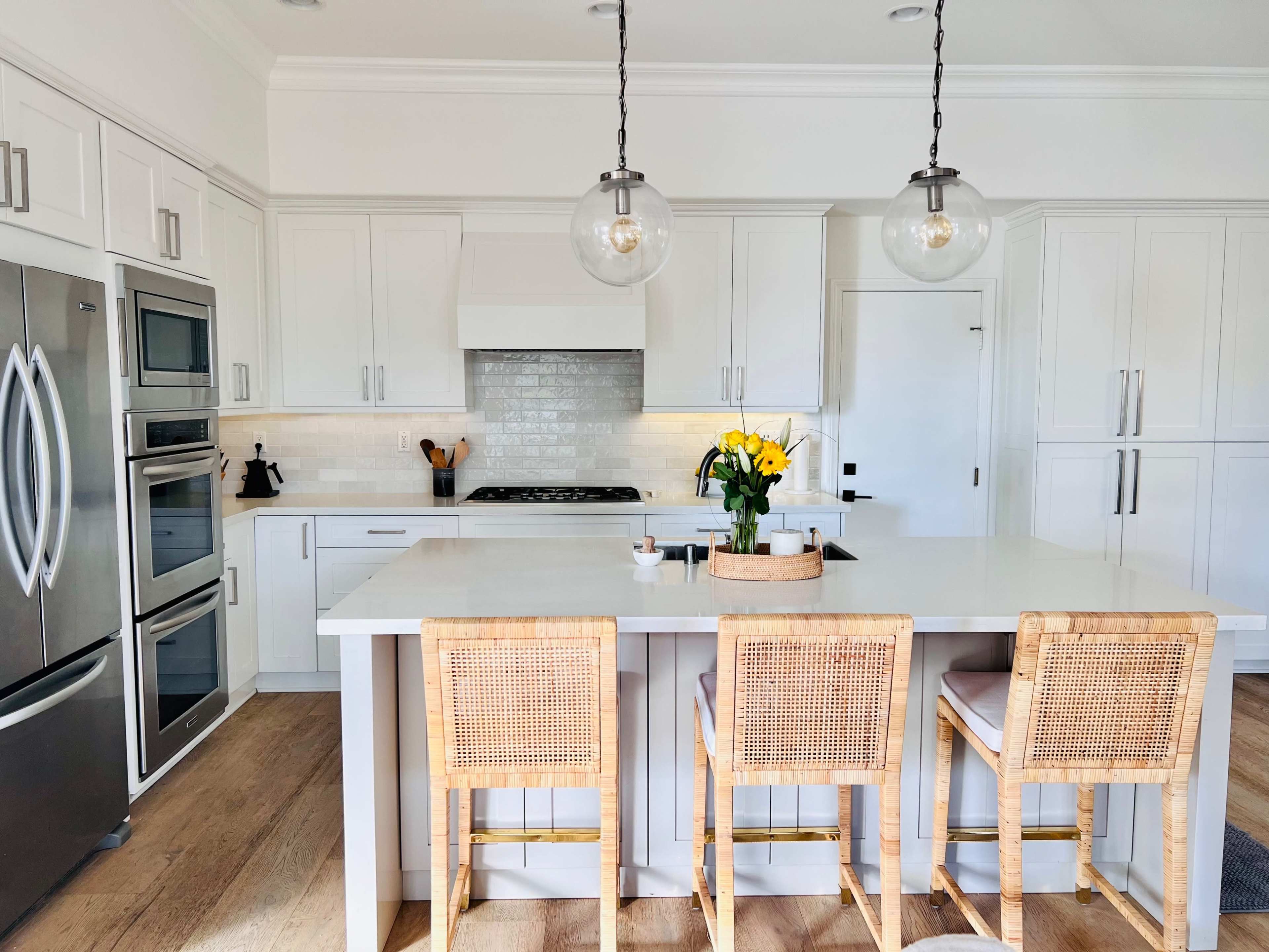 A modern kitchen features a central island with three rattan stools, stainless steel appliances, and a vase of yellow flowers on the countertop.