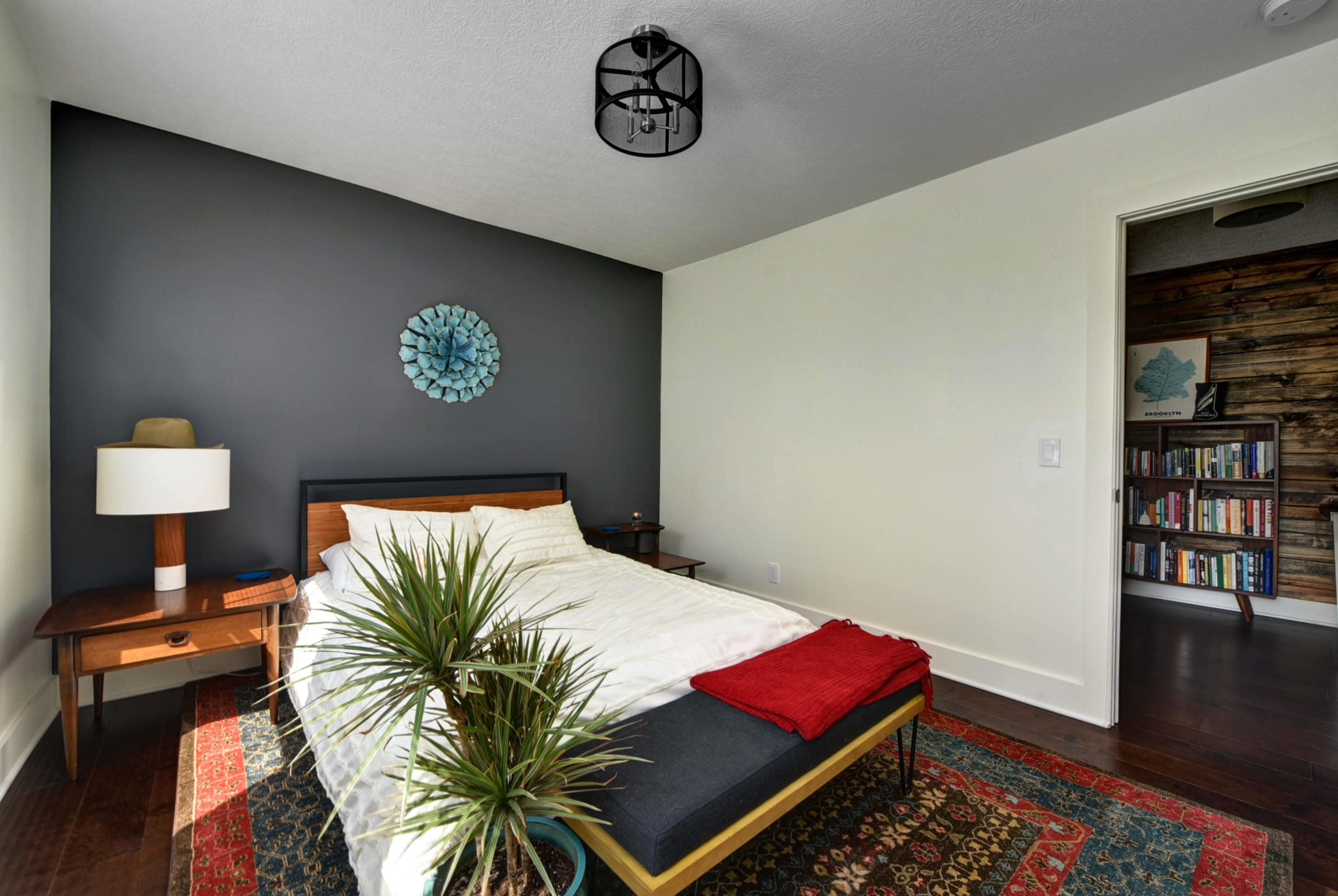 A minimalistic bedroom featuring a bed with a white comforter, a nightstand with a lamp, a decorative wall art piece, and a bookshelf visible in the background.
