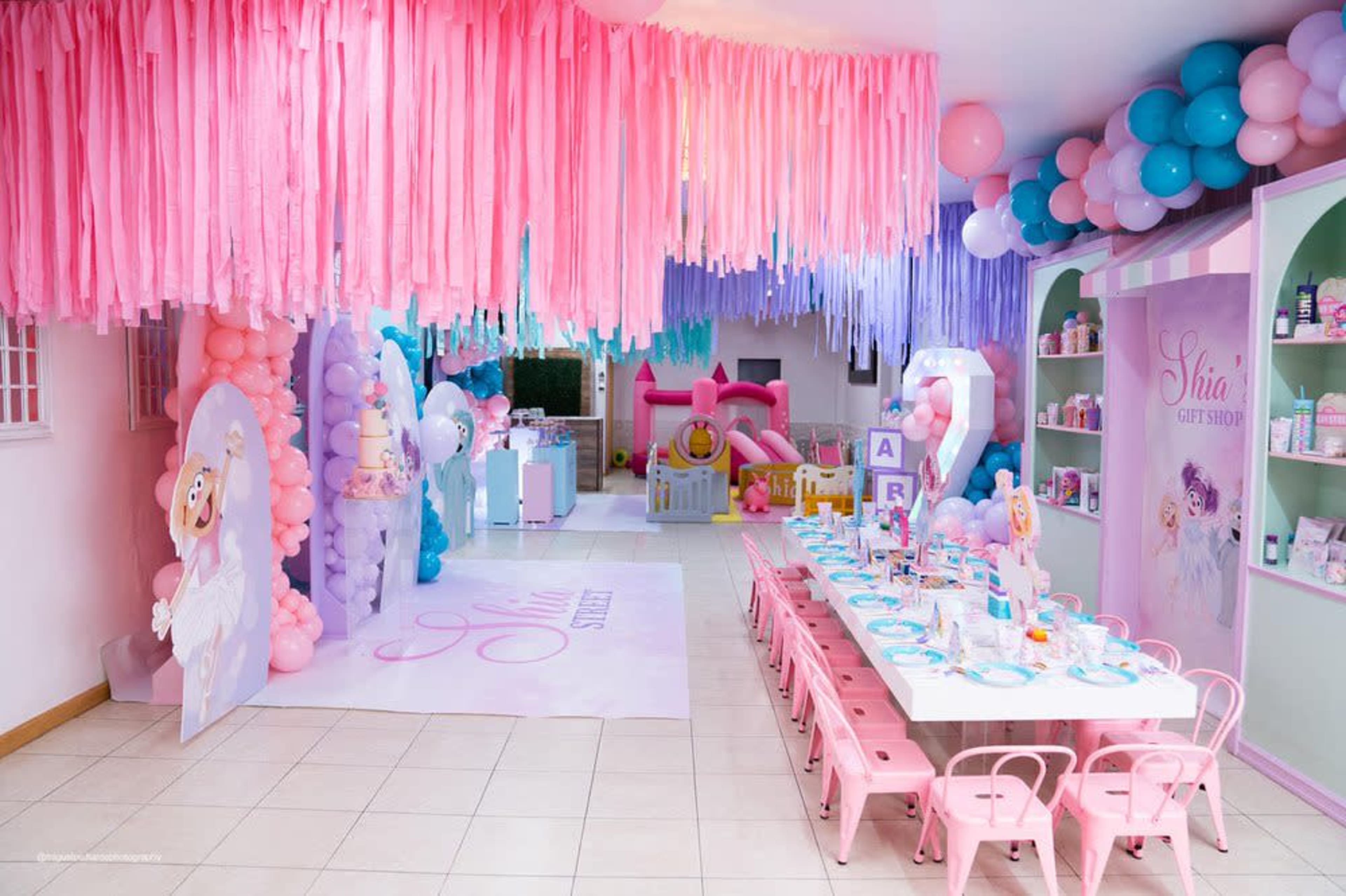 The image shows a brightly decorated party space featuring pink and purple streamers, balloon installations, a table set for children, and a play area with a small inflatable slide.