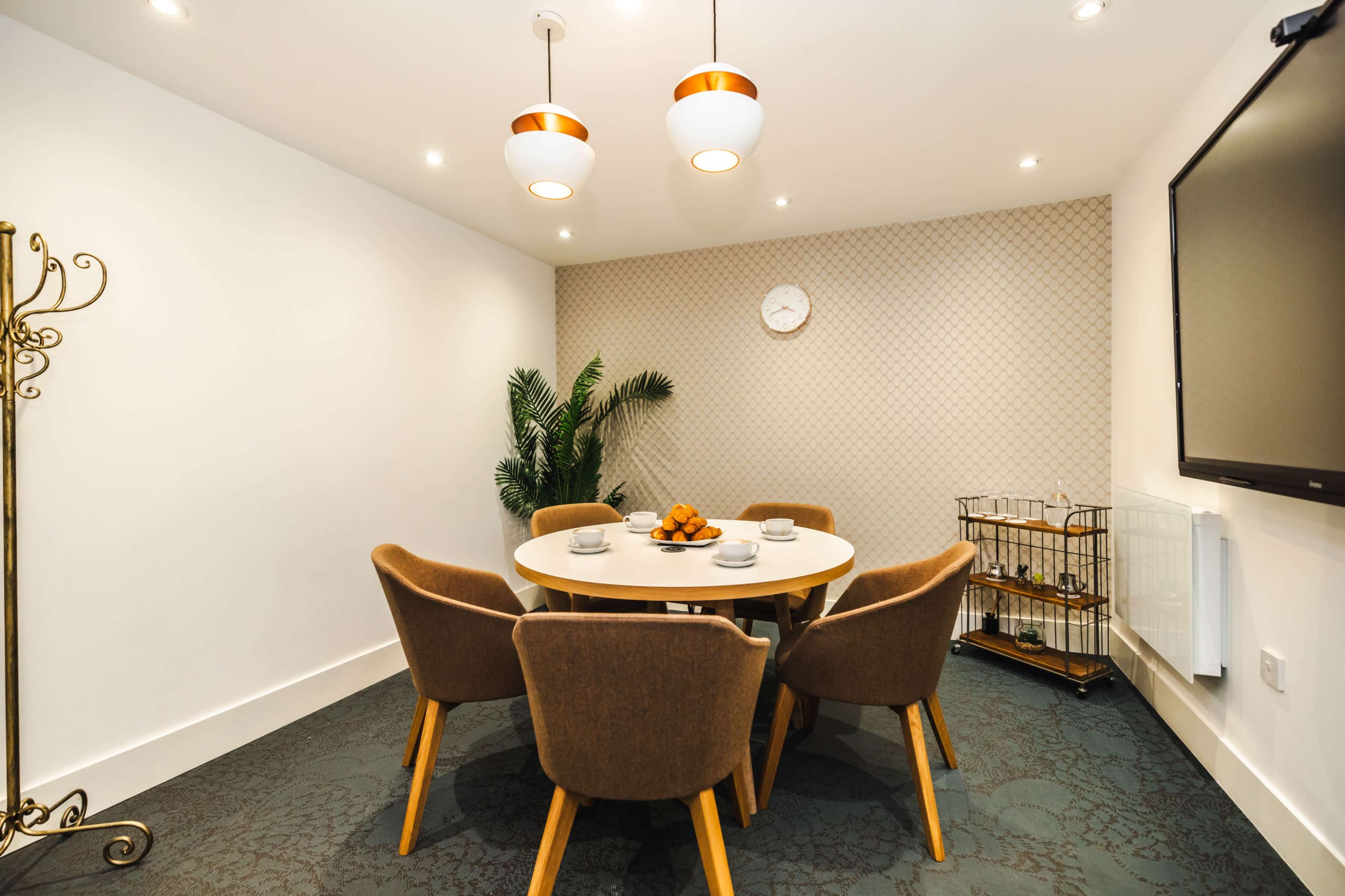 A round table with six chairs is set in a well-lit meeting room, featuring a wall clock and a decorative plant in a corner.