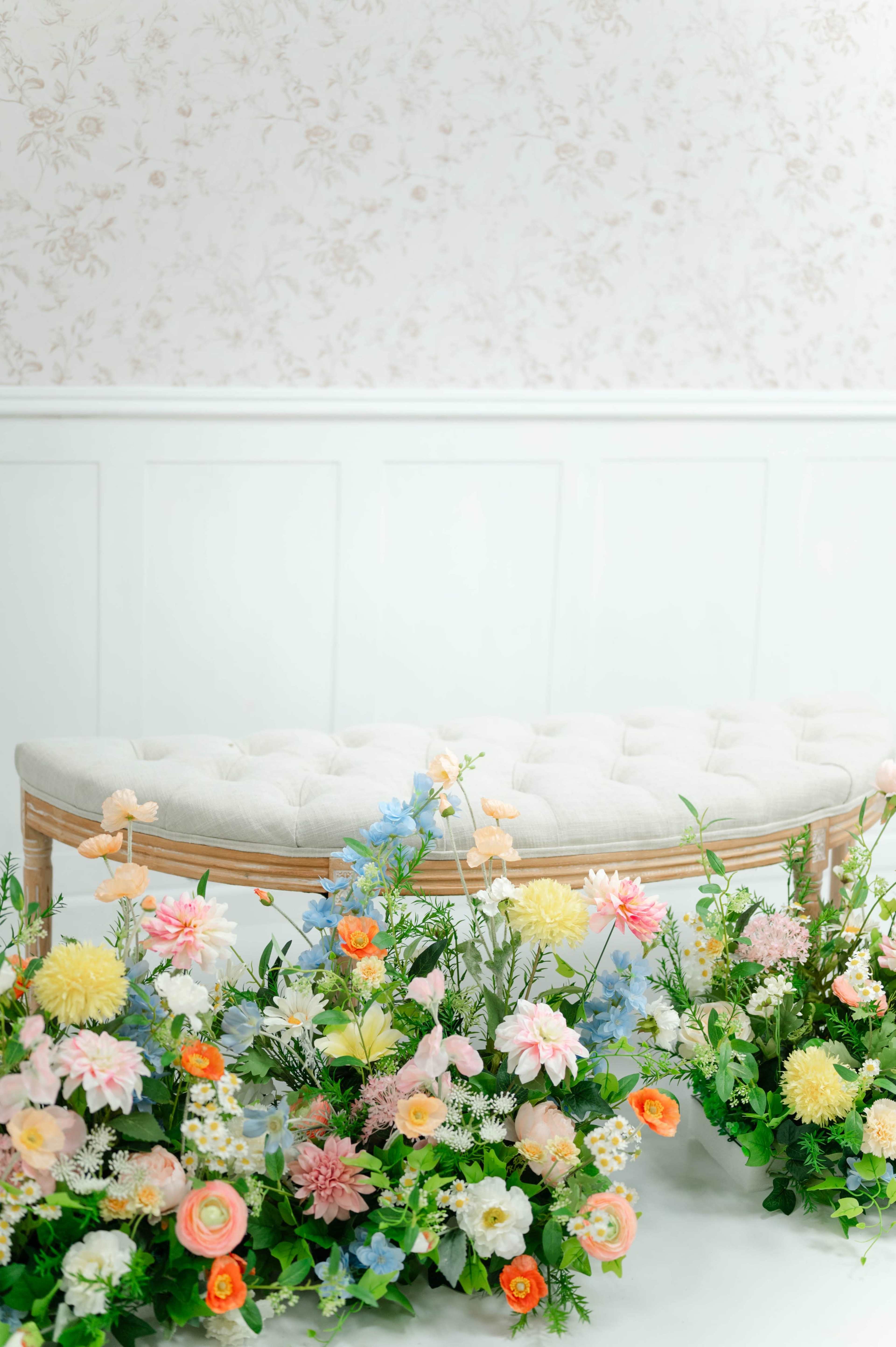 A tufted beige bench is positioned in front of a white wall adorned with floral wallpaper, surrounded by colorful flower arrangements.