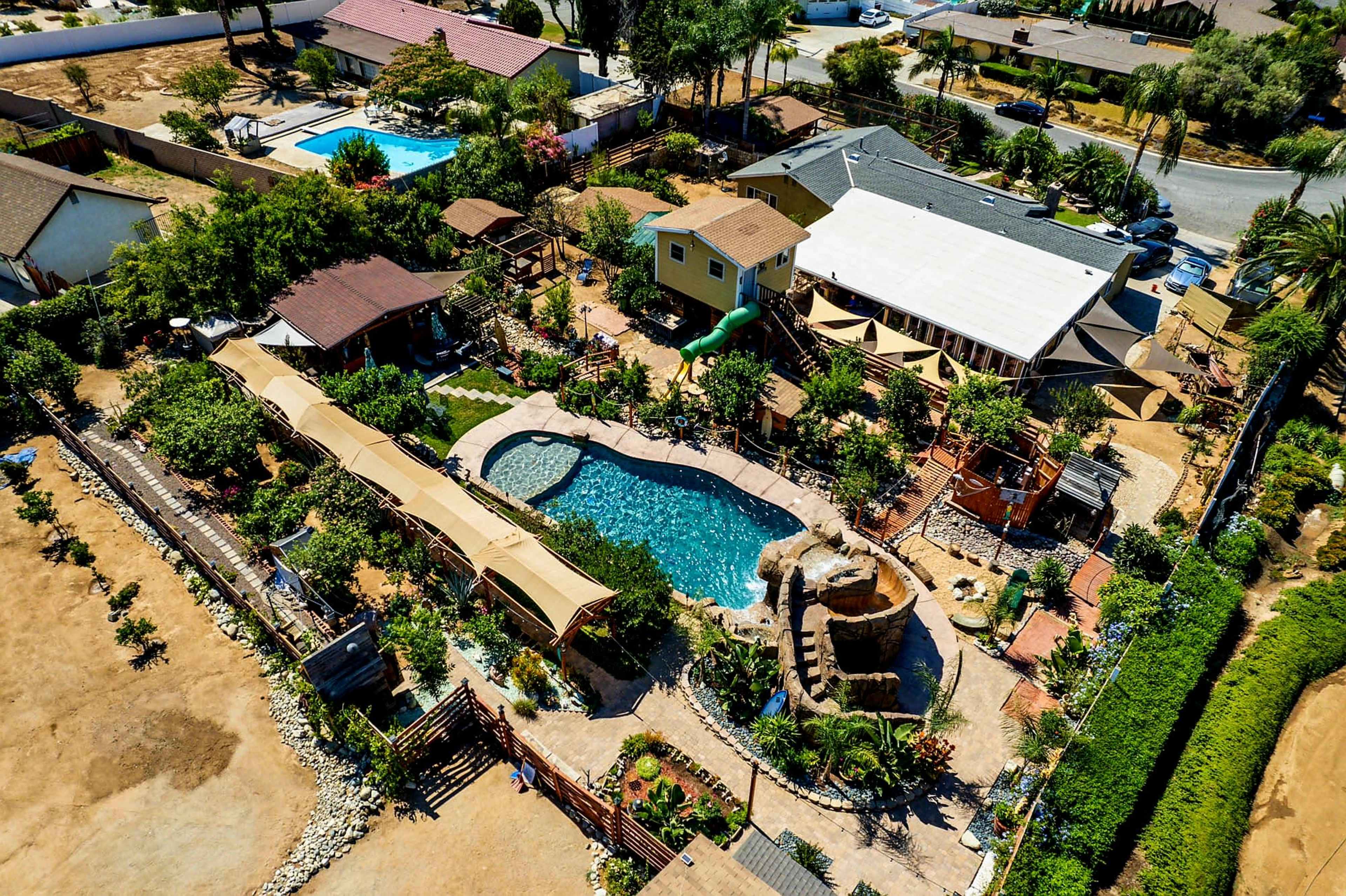 An aerial view shows a residential property with a swimming pool, landscaped gardens, and several structures, including pergolas and a water feature.