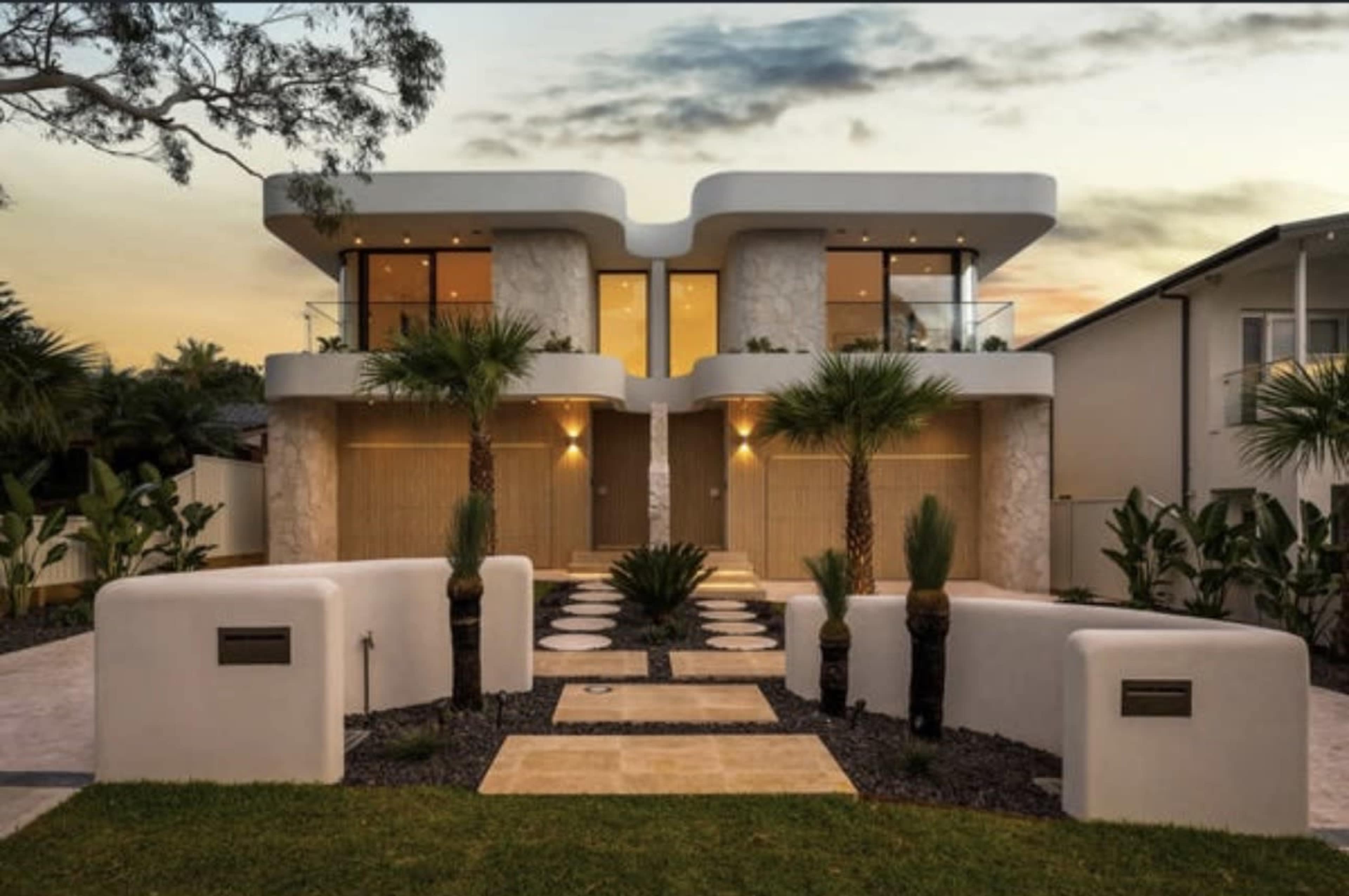 The image shows a modern, symmetrical house with two distinct entrances, surrounded by landscaped greenery and pathway stones.