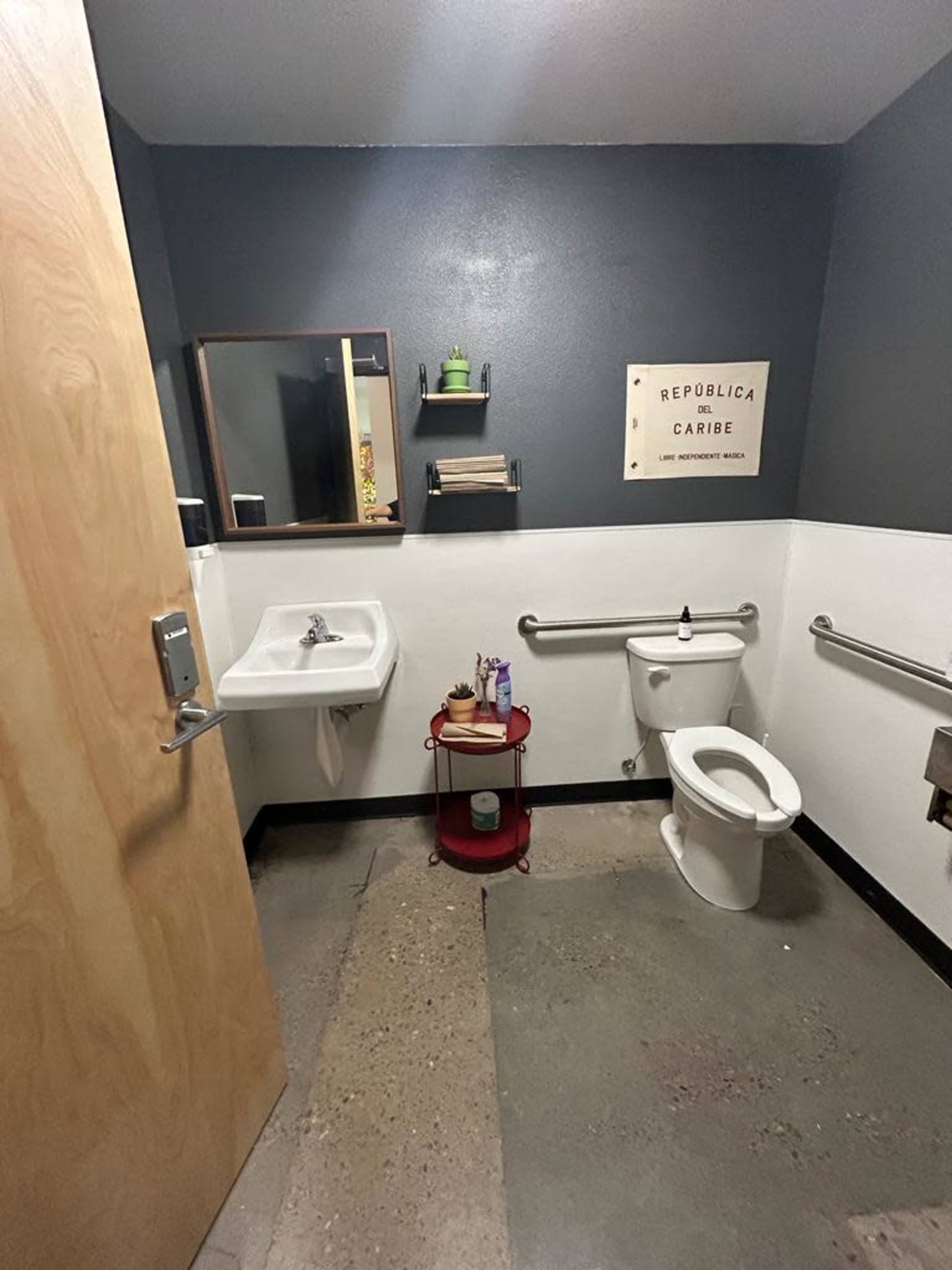 A small restroom with a sink, toilet, and a red cart holding various items, featuring gray walls and a wooden door.
