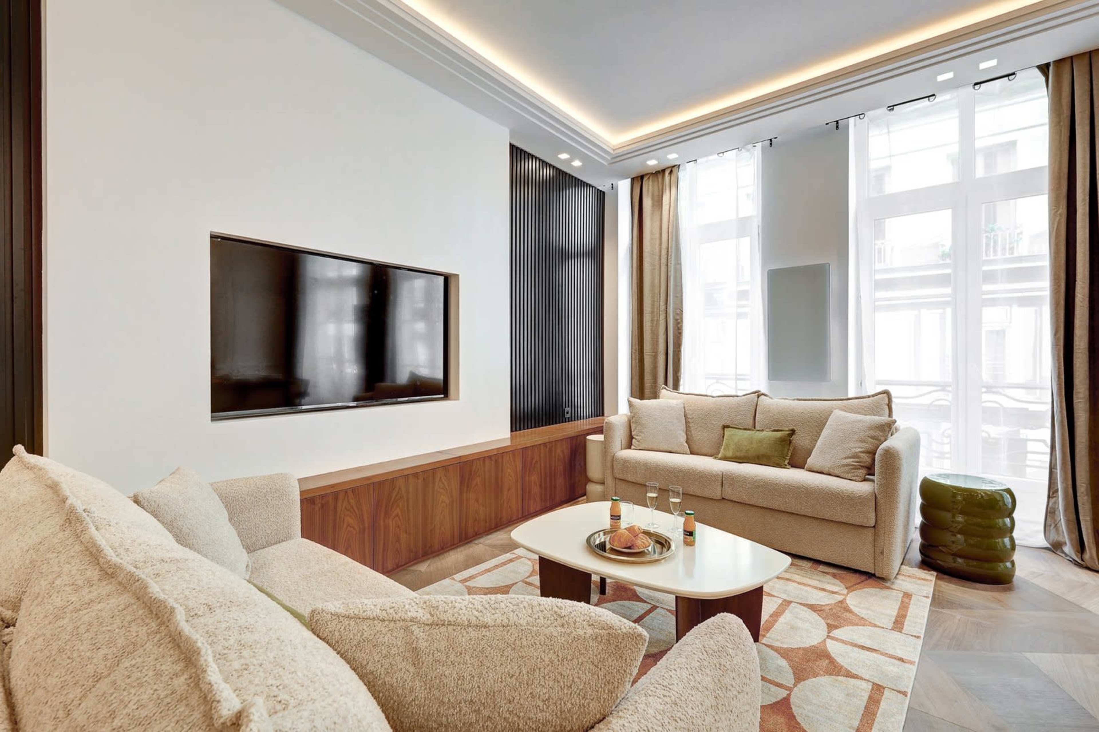 A modern living room features two sofas, a round coffee table, and a wall-mounted television, with large windows allowing natural light to enter the space.