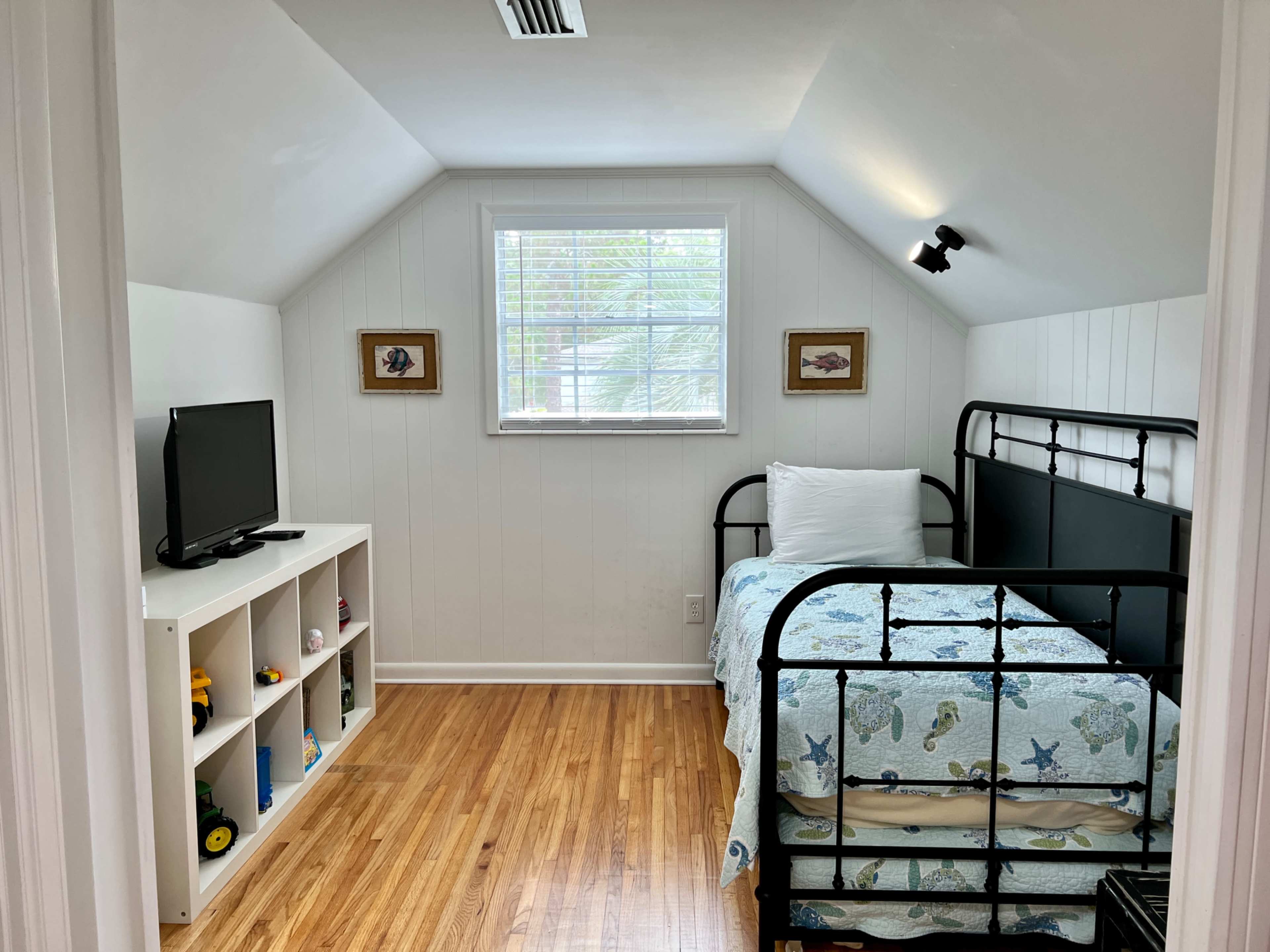 A small, tidy bedroom with a bed covered in a light blue patterned blanket, a television on a white shelf, and a window allowing natural light.