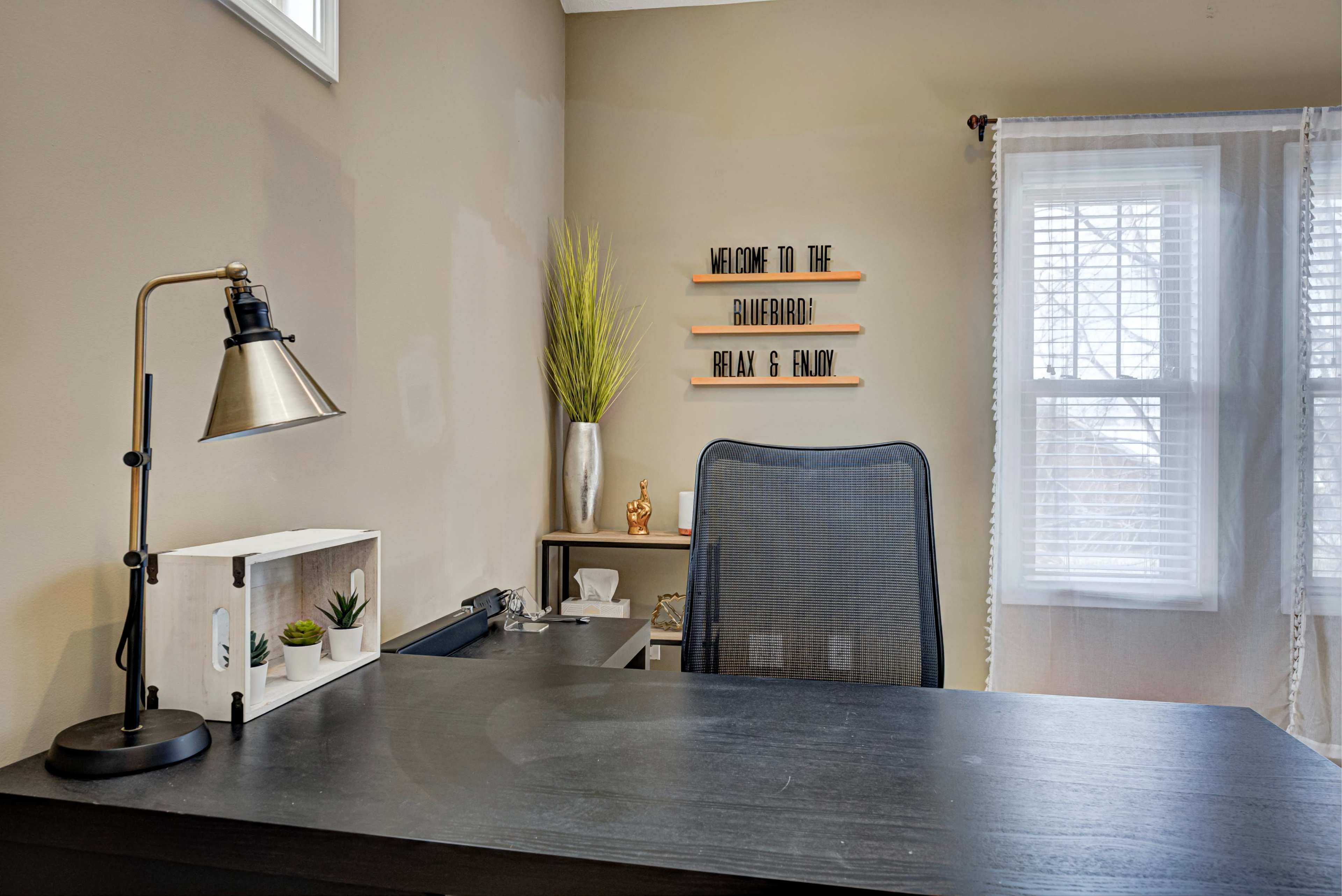 A modern office space features a black desk with a lamp, a mesh chair, and decorative elements including a plant and a welcome sign on the wall.