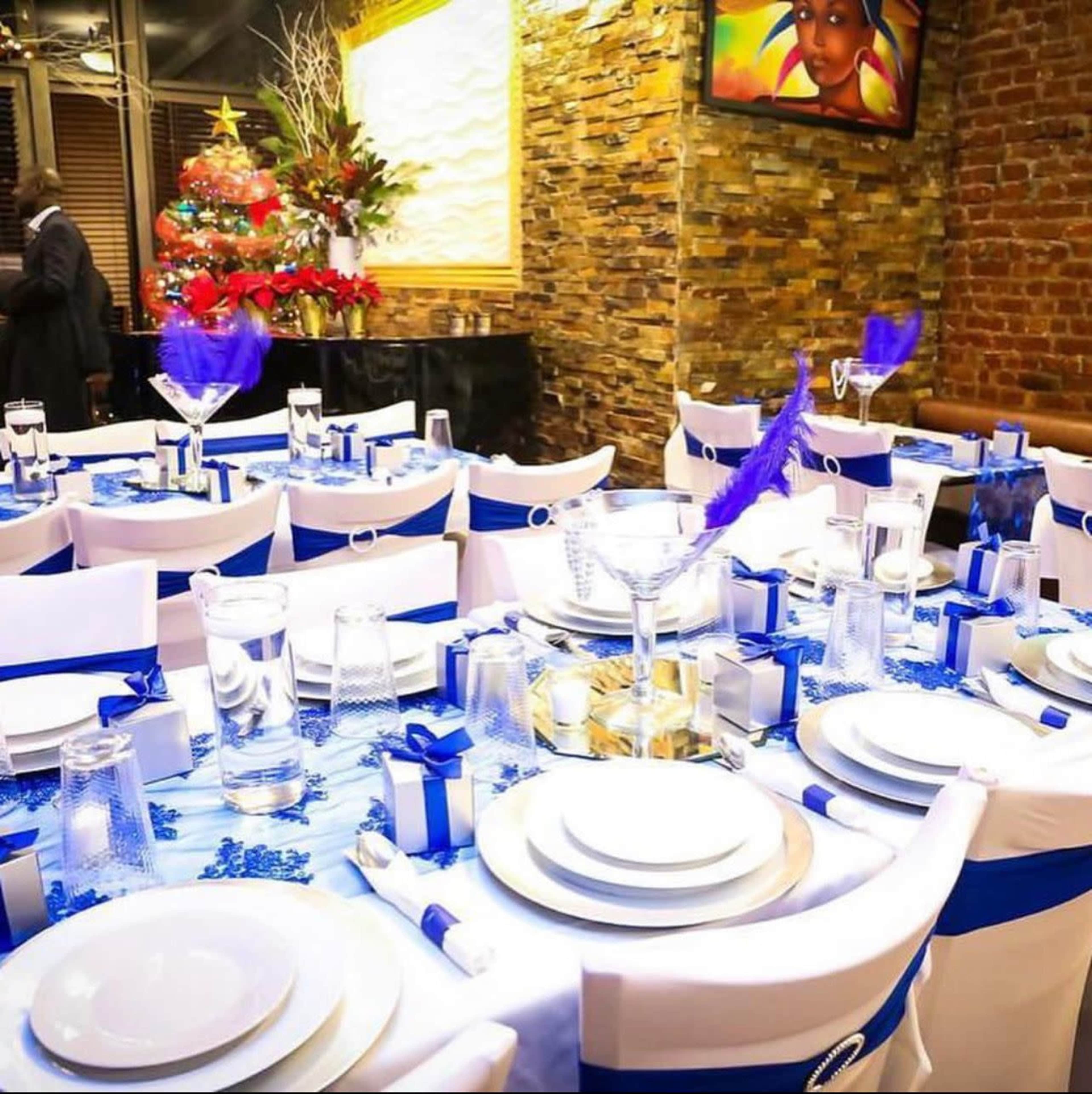 A banquet hall is set up with elegantly arranged tables featuring white and blue tablecloths, decorative centerpieces, and glassware, alongside a Christmas tree in the background.