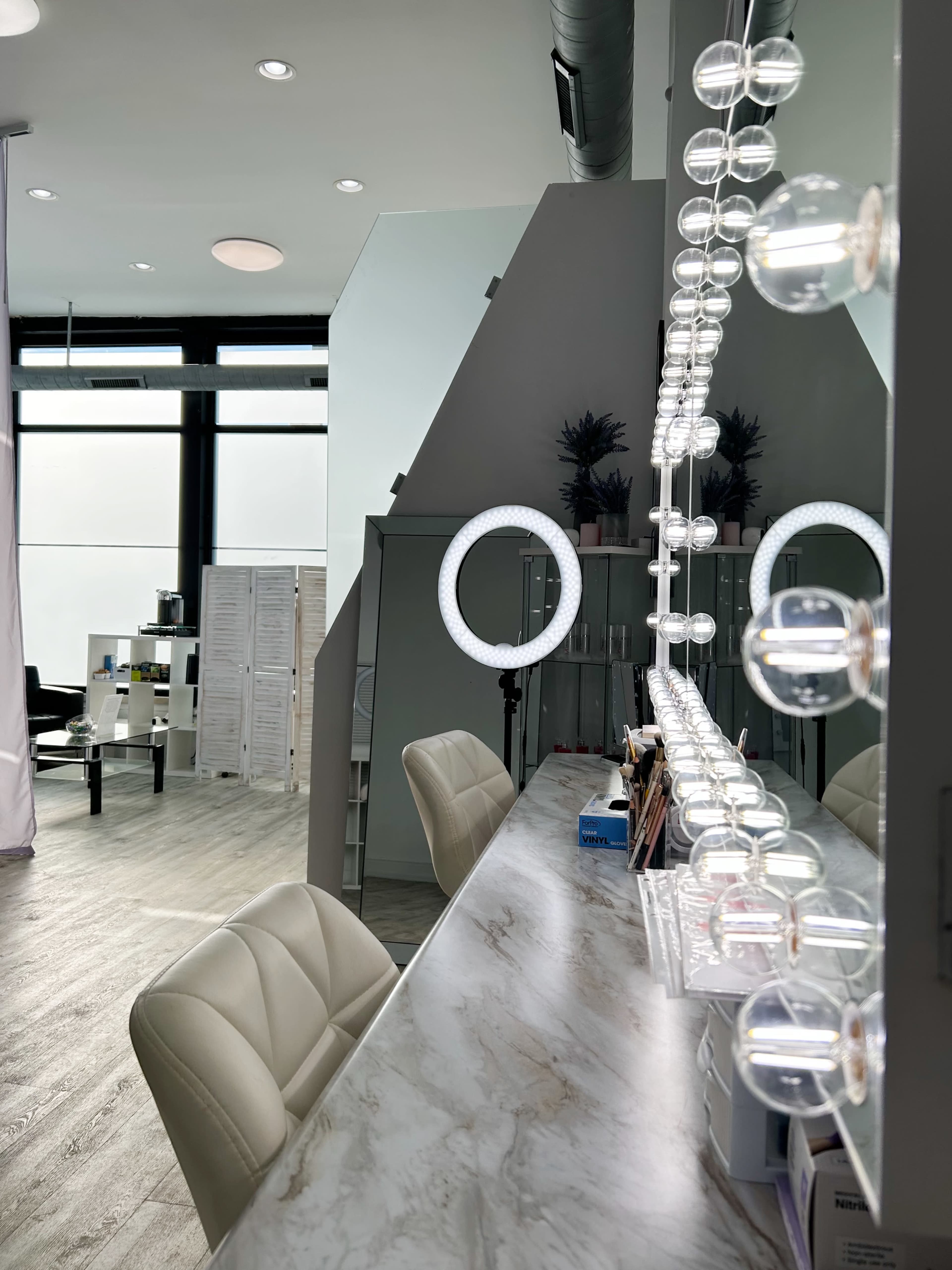A modern beauty salon interior with a large makeup mirror illuminated by round bulbs, and a sleek marble counter.