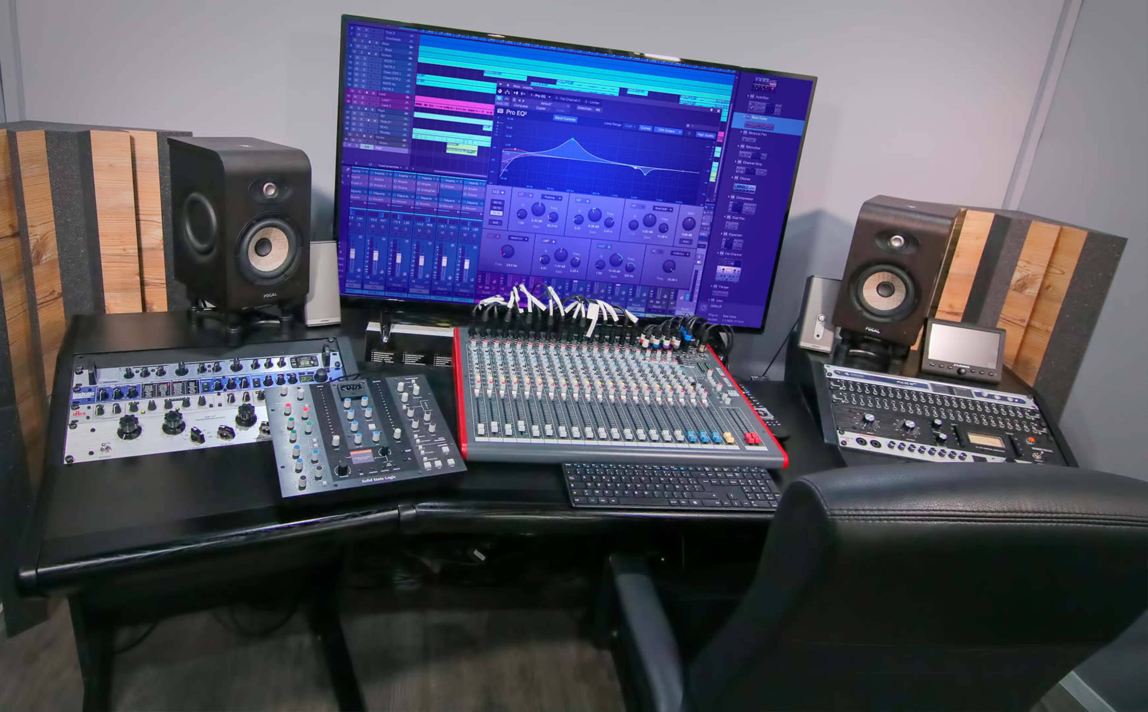A music production setup with a large monitor displaying audio software, a mixing console, audio interfaces, and studio monitors arranged on a desk.