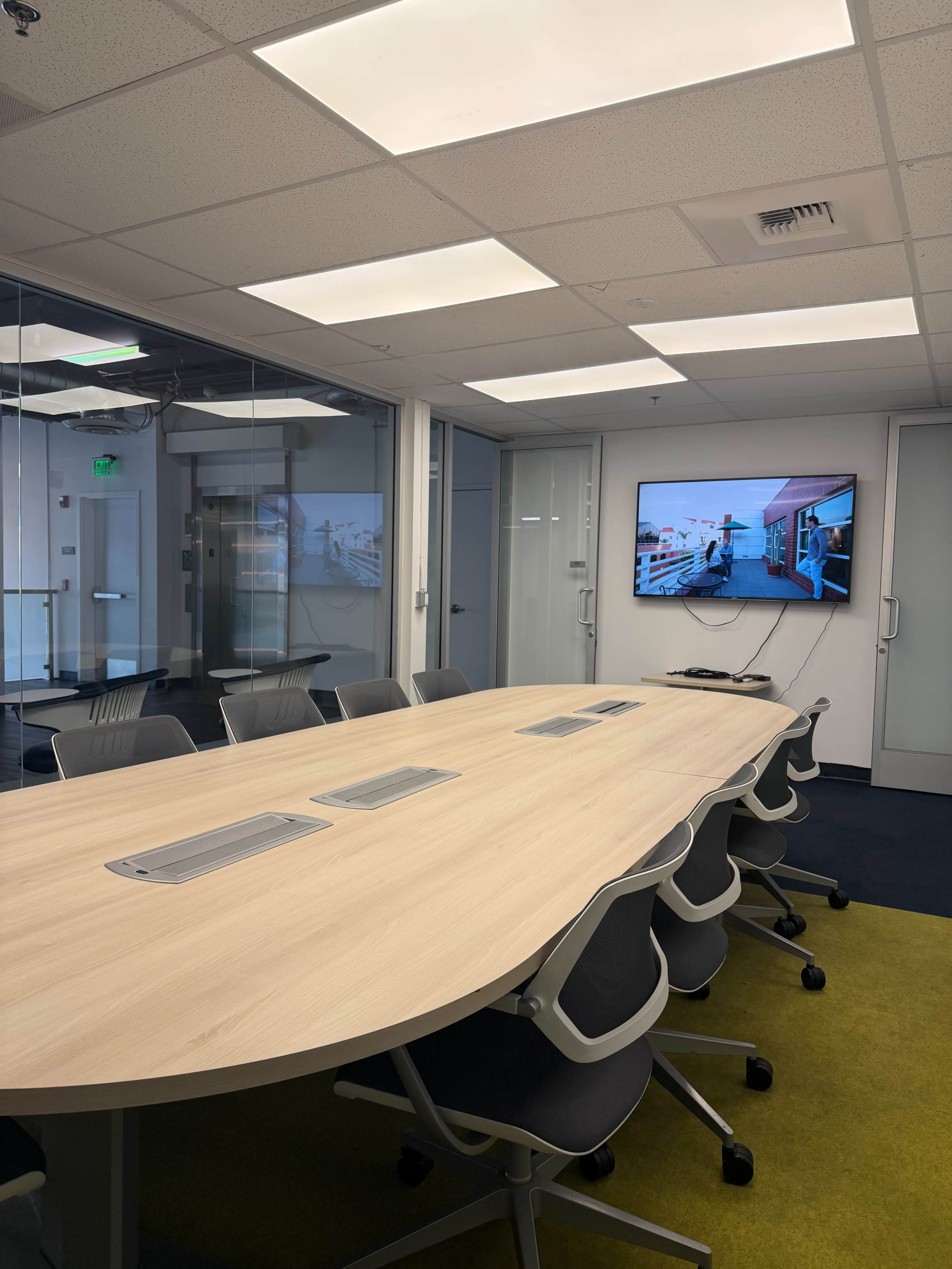 A modern conference room features a large oval table surrounded by gray chairs, with a screen displaying visuals in the background.
