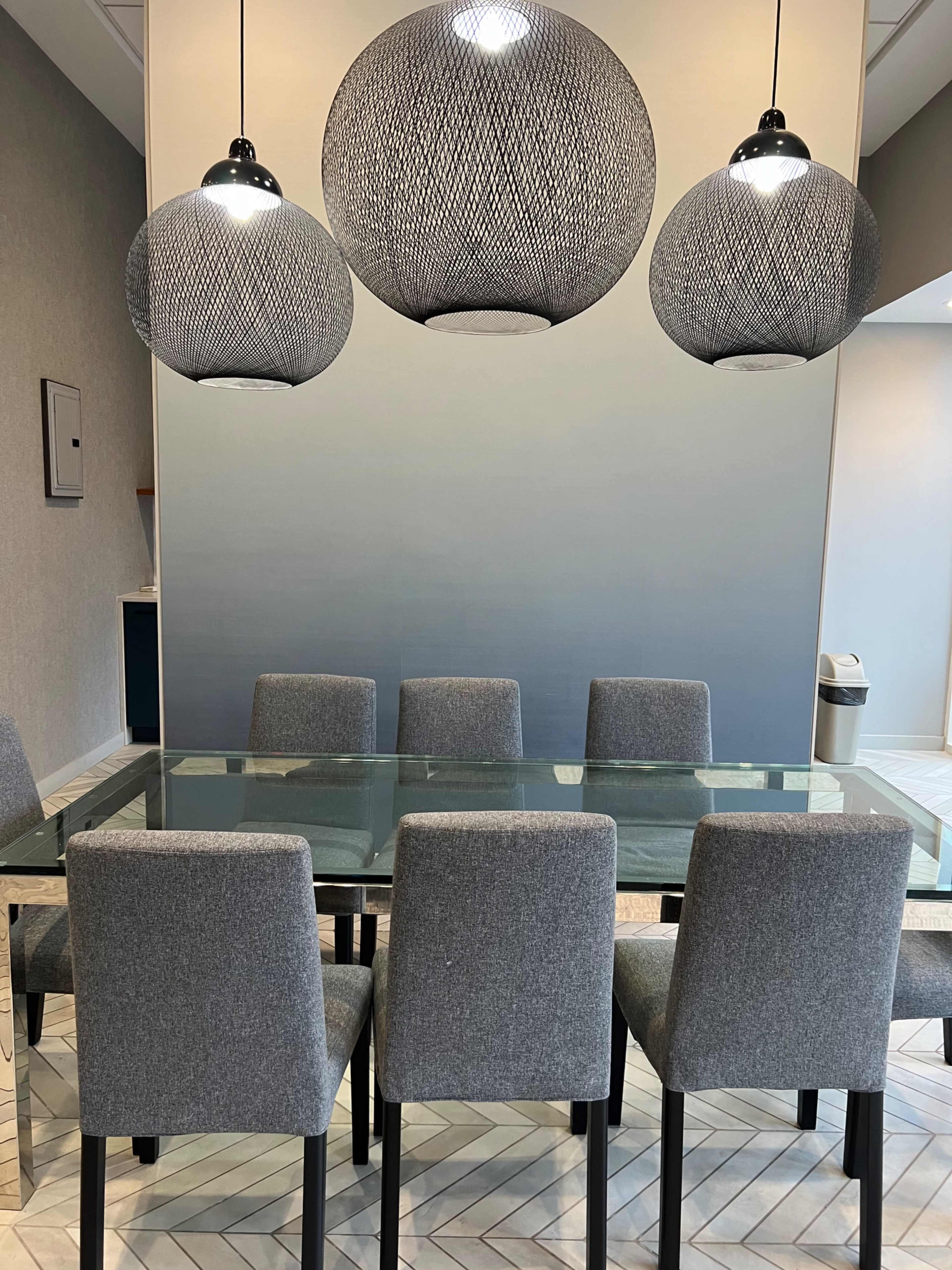 A modern dining area features a glass table surrounded by four gray upholstered chairs, with three black pendant lights hanging above.