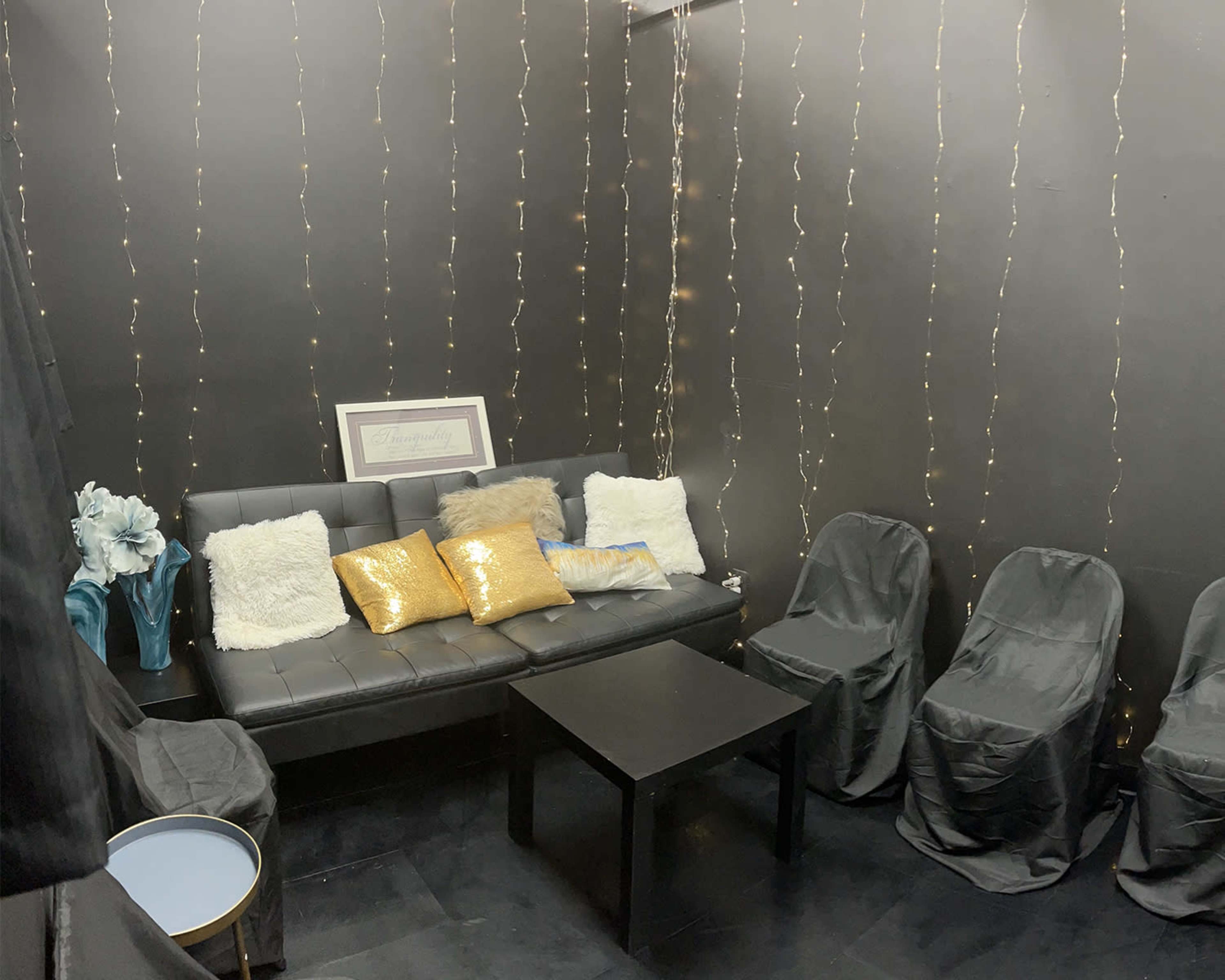 A small black-themed lounge area with a black sofa adorned with colorful cushions, a coffee table, and several chairs draped in black fabric, alongside fairy lights on the walls.