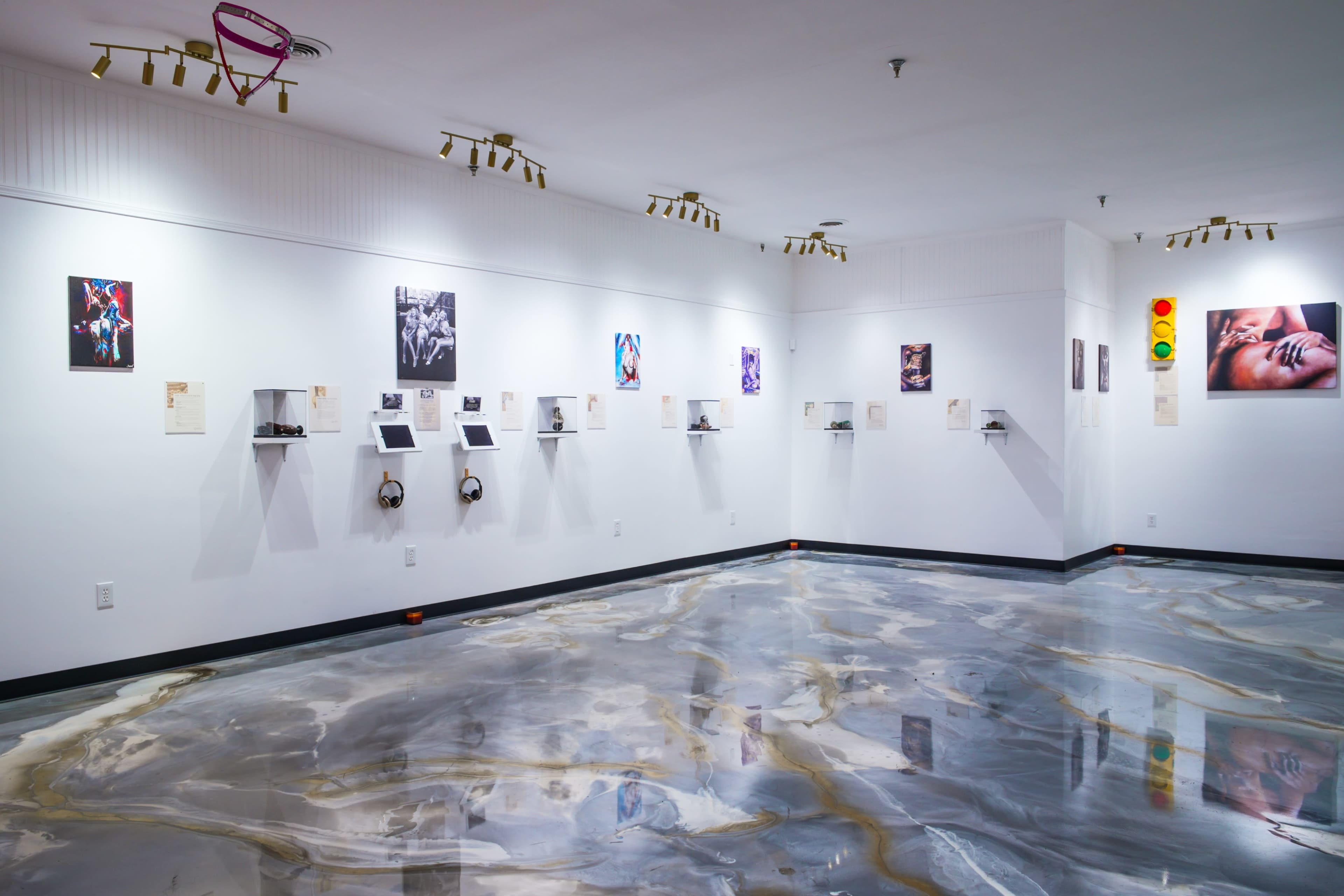 The image shows a modern art gallery with a polished floor and several artworks displayed on the white walls, along with audio equipment mounted on shelves.