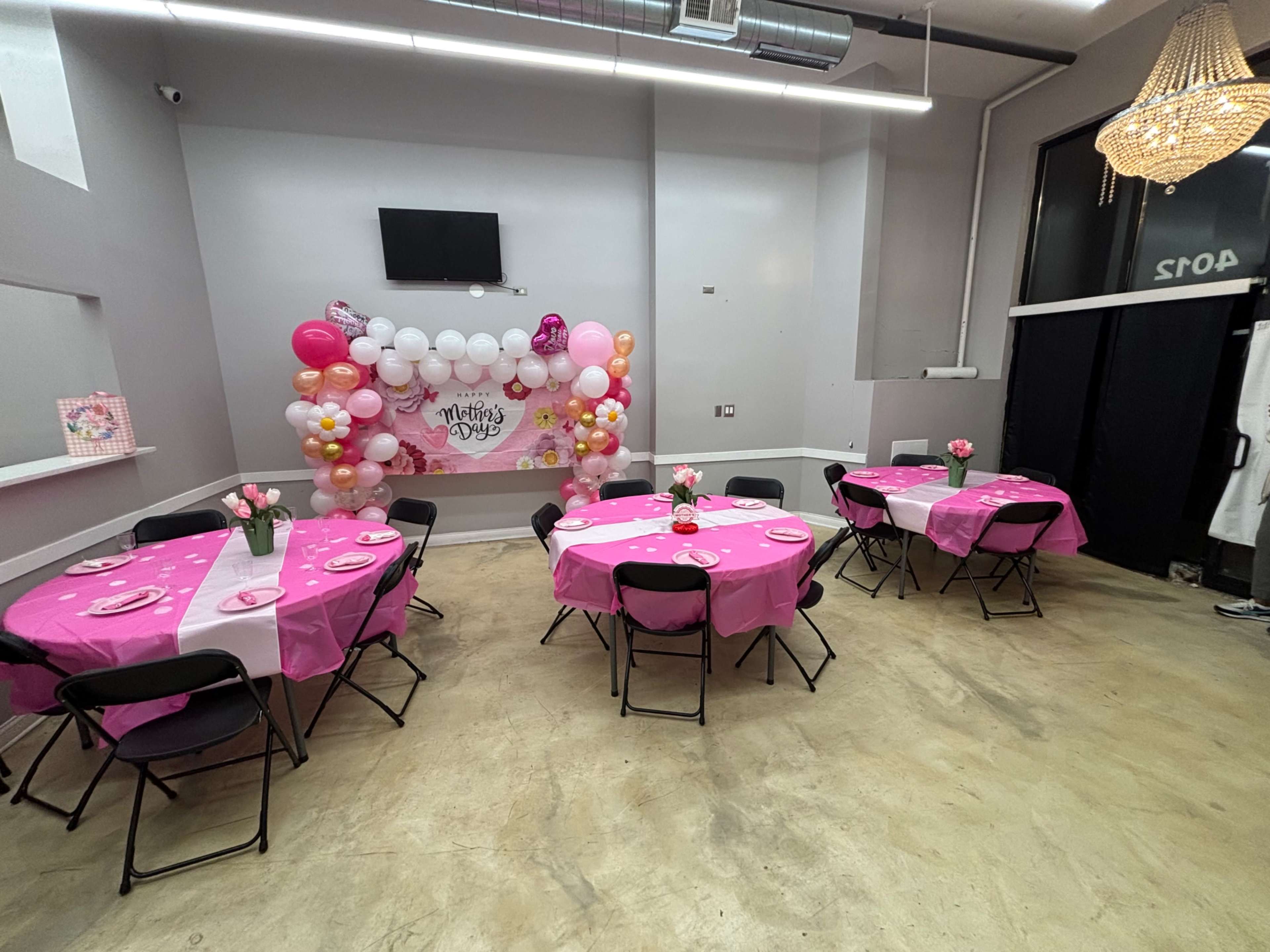 The image shows a party room set up with pink tablecloths and decorations, featuring tables arranged around a colorful balloon backdrop.