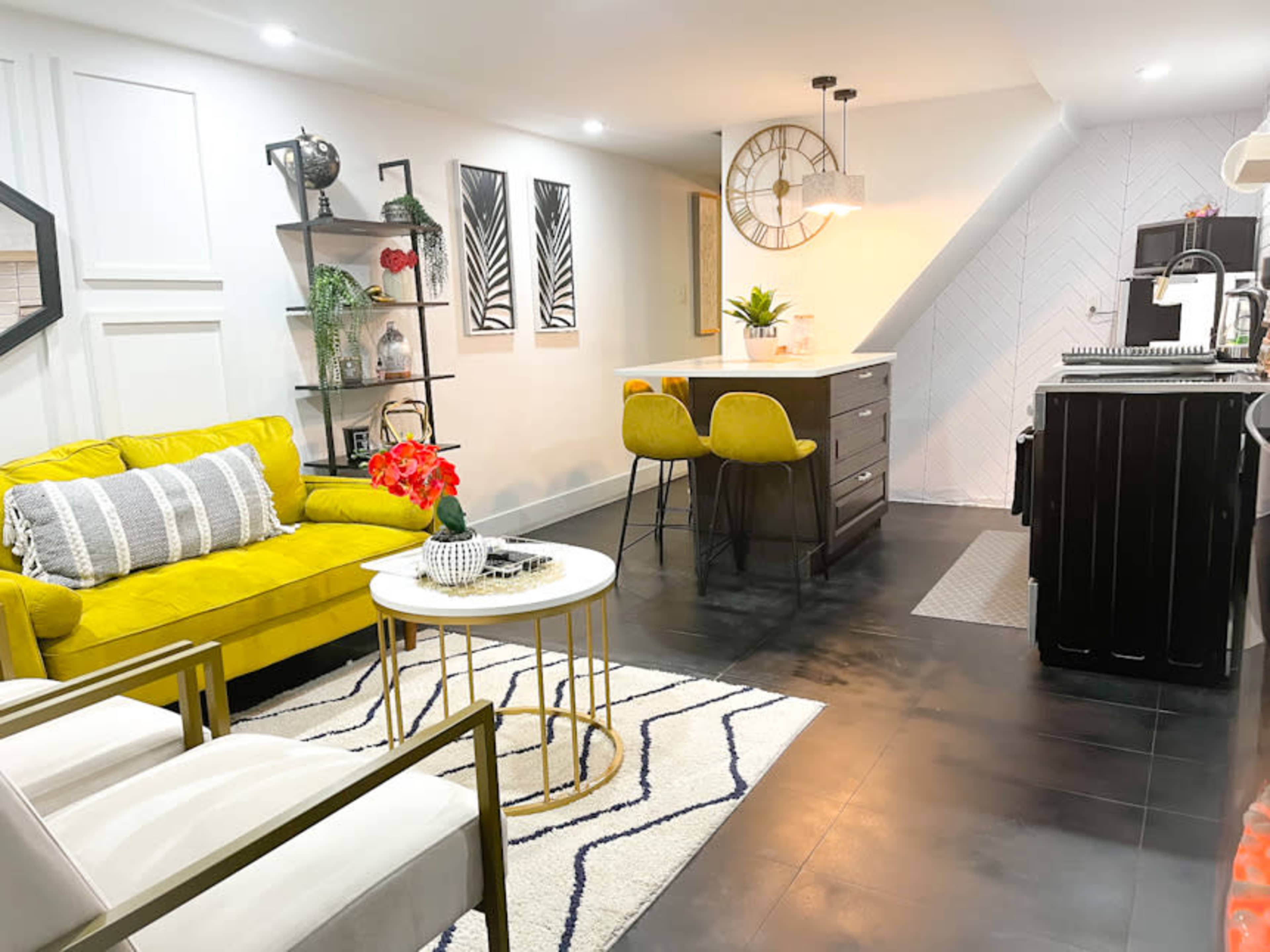A modern living space features a yellow sofa, a circular coffee table, and a kitchen area with bar stools and contemporary decor.