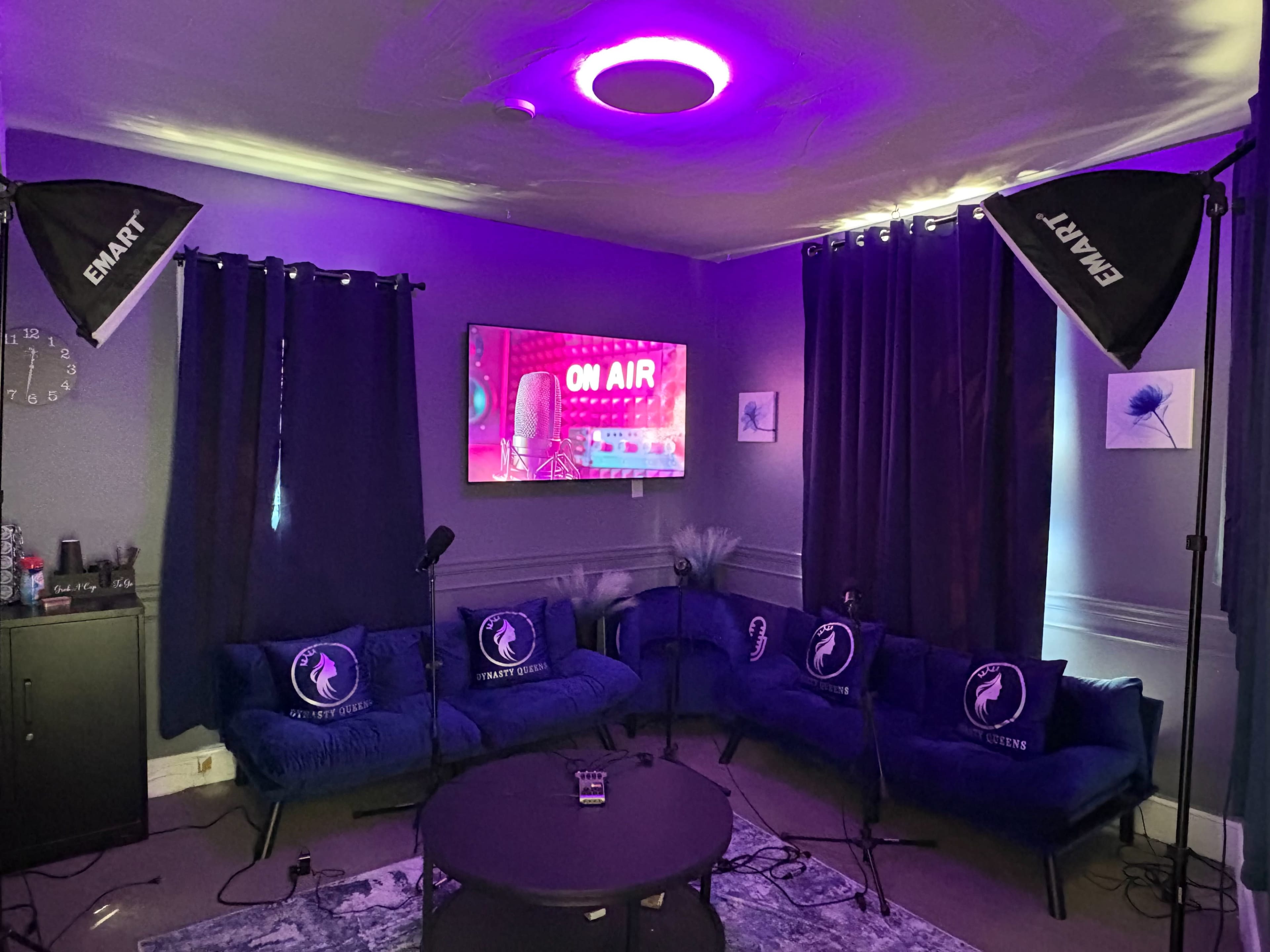 A modern lounge area features blue couches with decorative pillows, a large television displaying "ON AIR," and professional lighting setups.