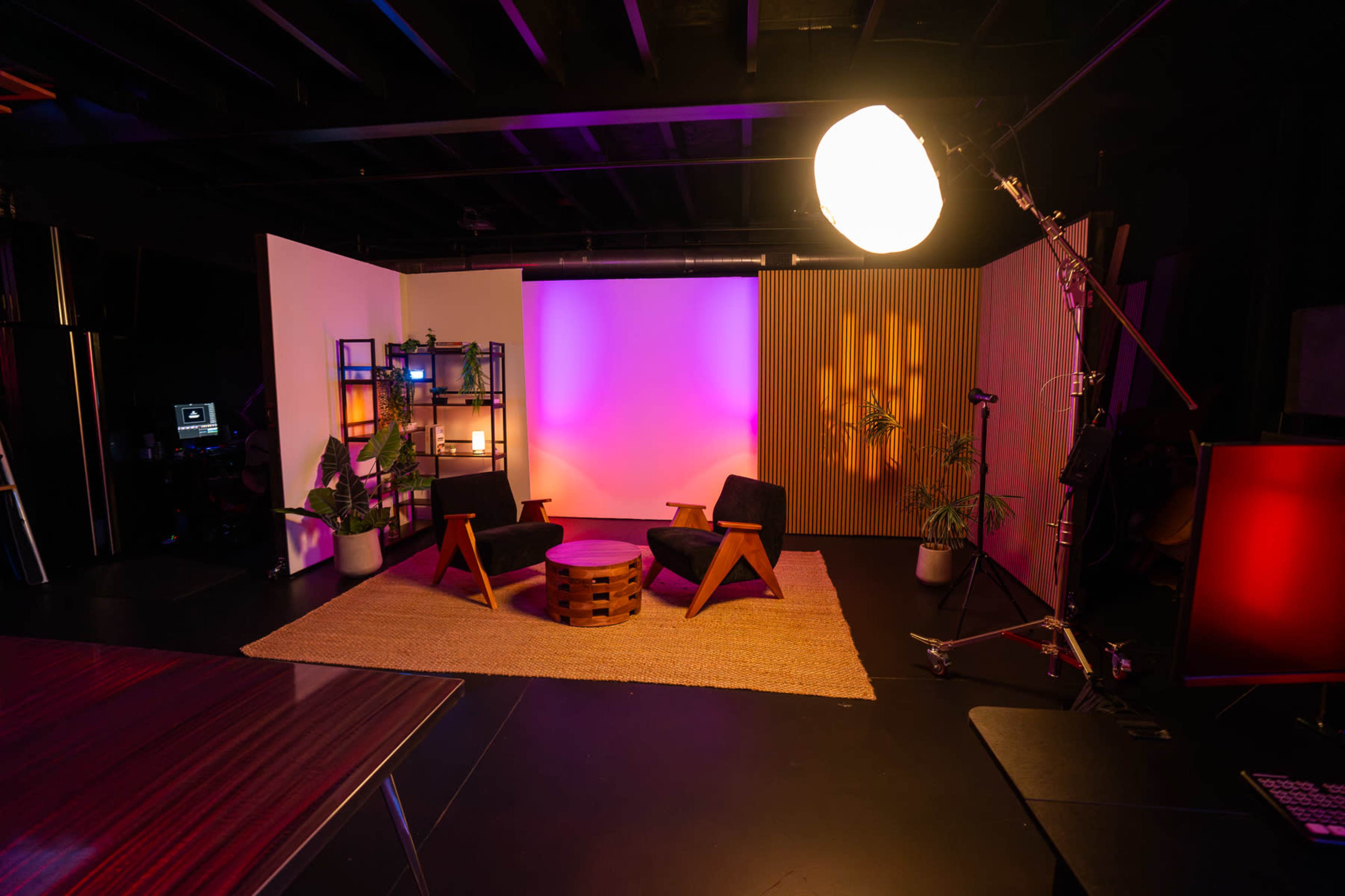 The image shows a modern studio setup featuring two black chairs, a round wooden table, and decorative plants, all surrounded by colorful lighting and various equipment.