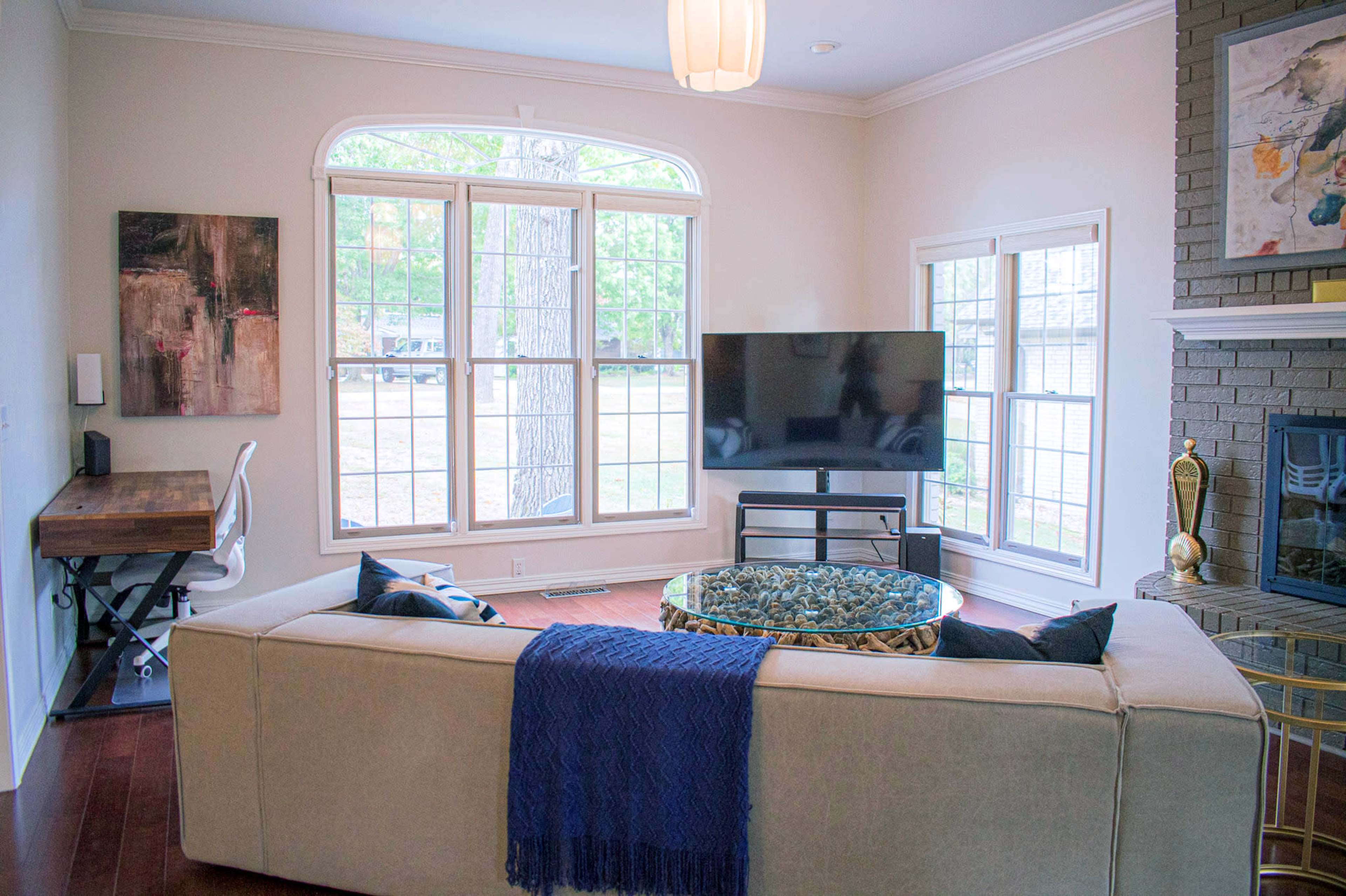 A living room features a beige sofa with a blue throw, a glass coffee table, a television on a stand, and large windows overlooking a garden.