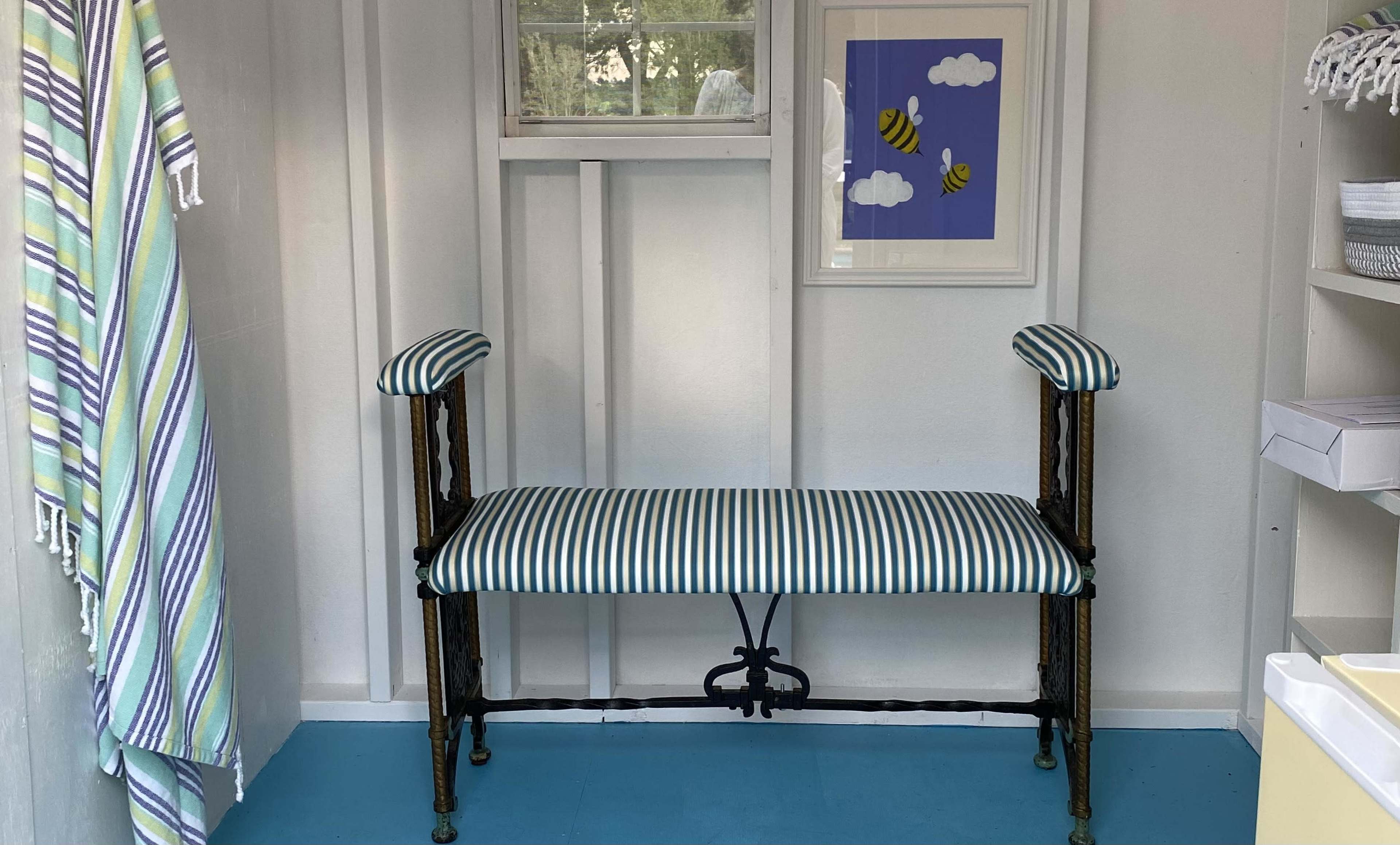 A room with a striped upholstered bench, a window, and a framed artwork featuring clouds and bees on the wall.