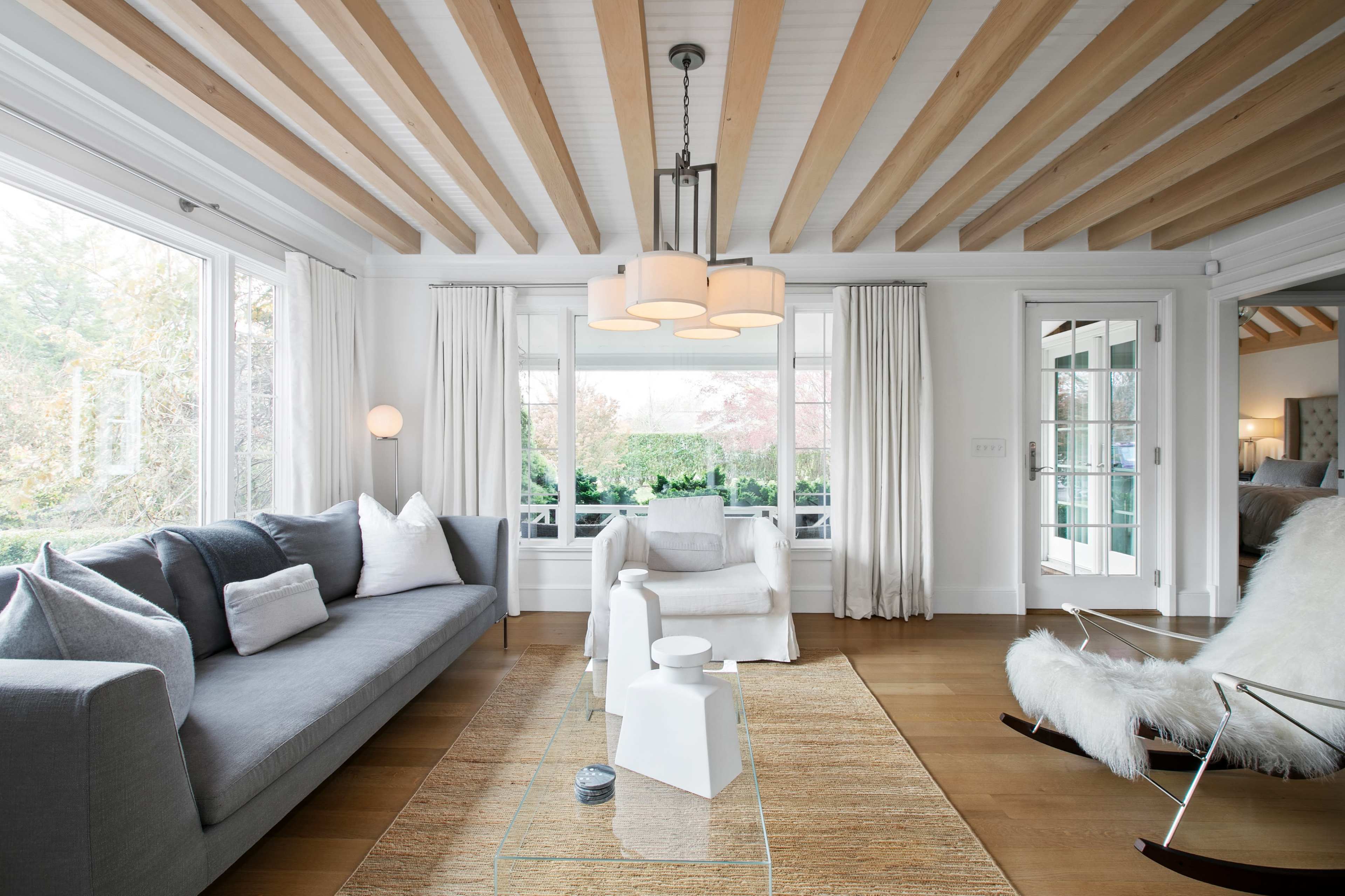 A well-lit living room features a gray sofa, a white chair, a glass coffee table, and wooden beamed ceilings with large windows overlooking a garden.