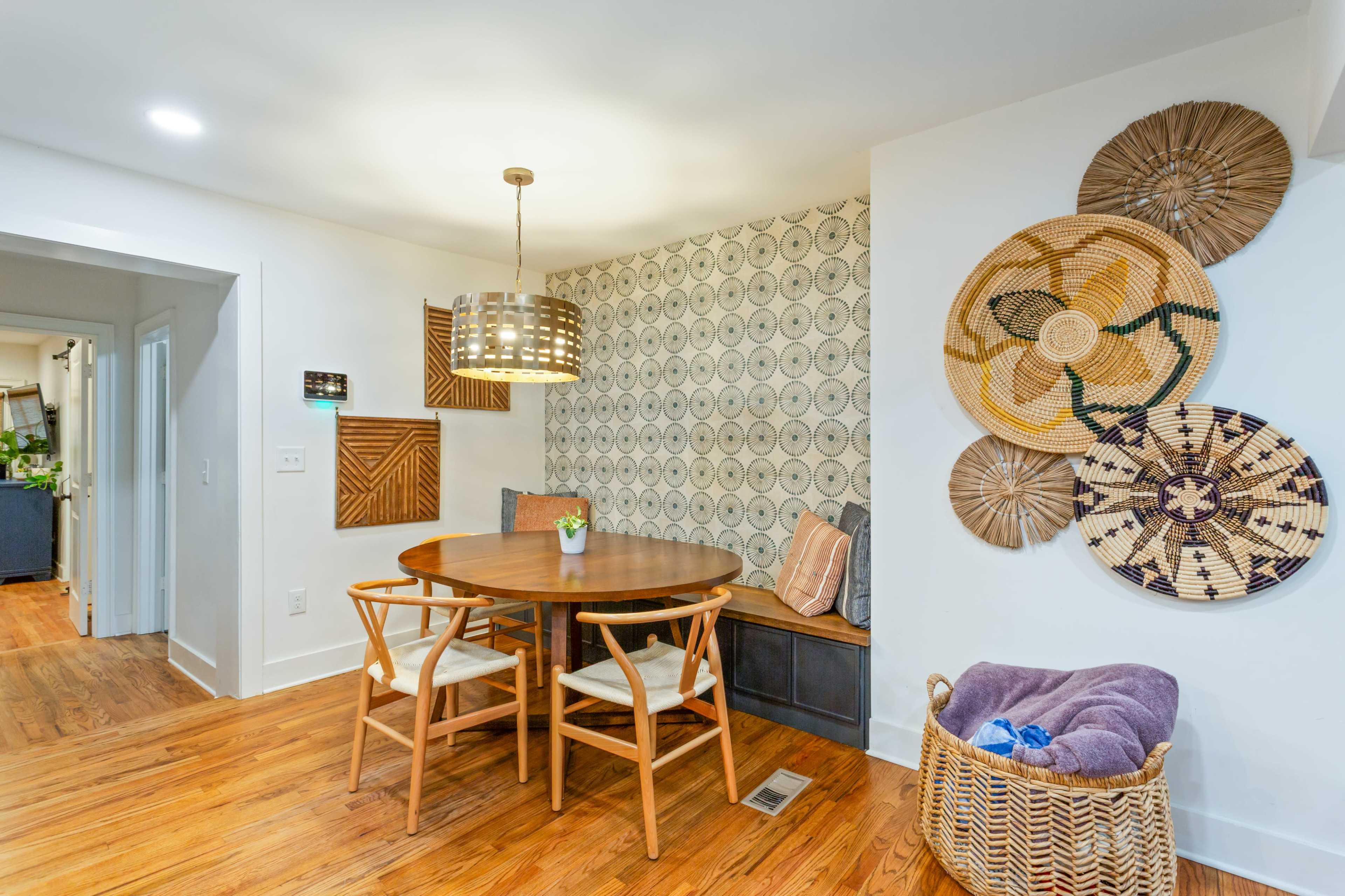 The image shows a dining area featuring a round wooden table surrounded by white chairs, with a decorative wall adorned with woven baskets and a patterned wallpaper.