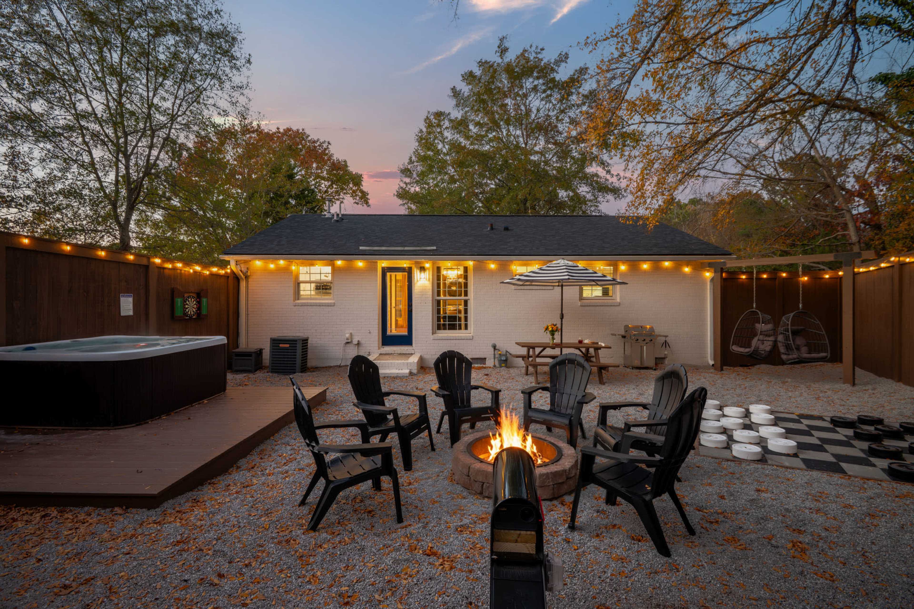 The image shows a backyard with a fire pit surrounded by black chairs, a hot tub on the left, and a white house with string lights and a table set up in the center.