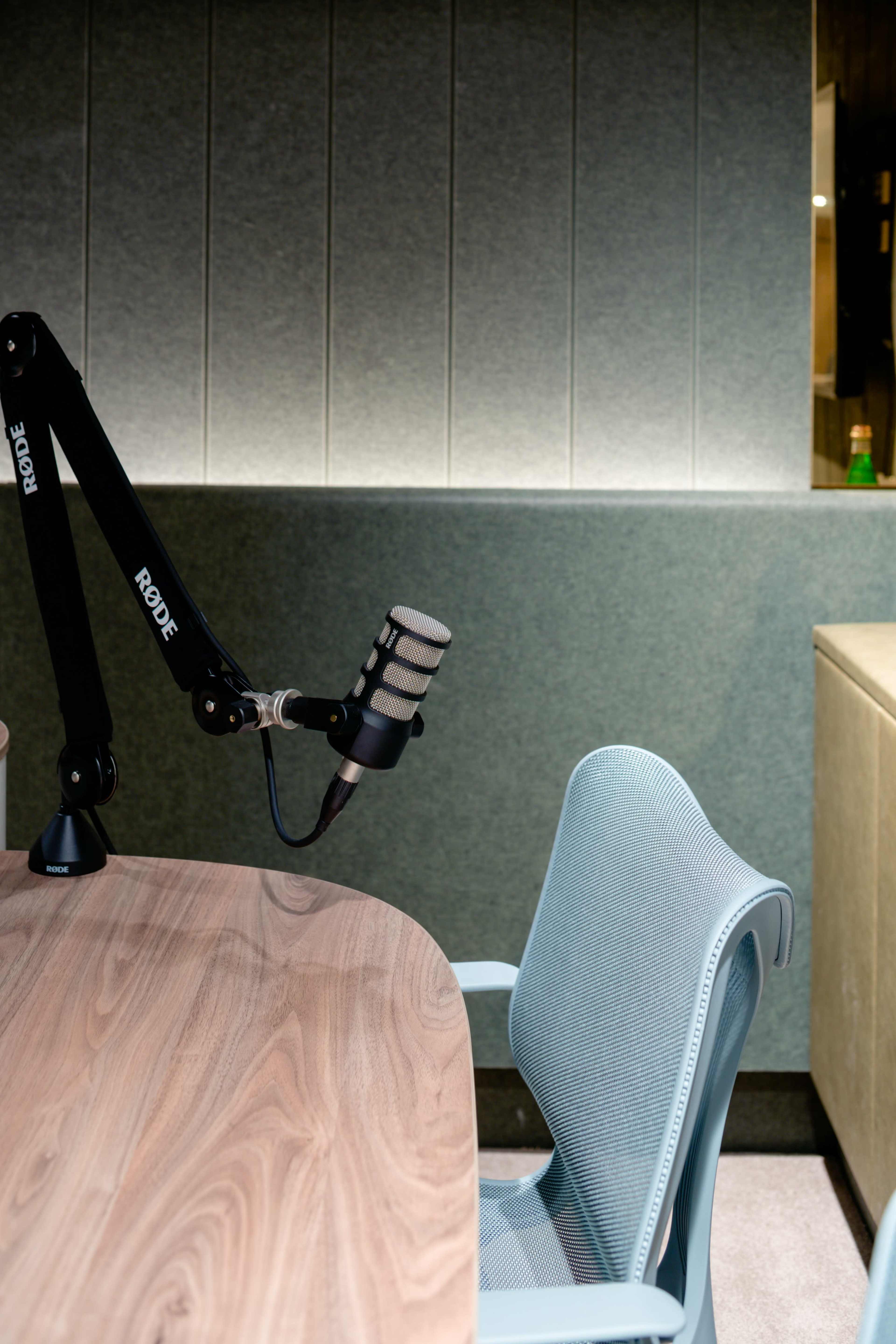 A Rode microphone is mounted on a boom arm next to a modern wooden desk and a light blue ergonomic chair in a softly lit studio setting.