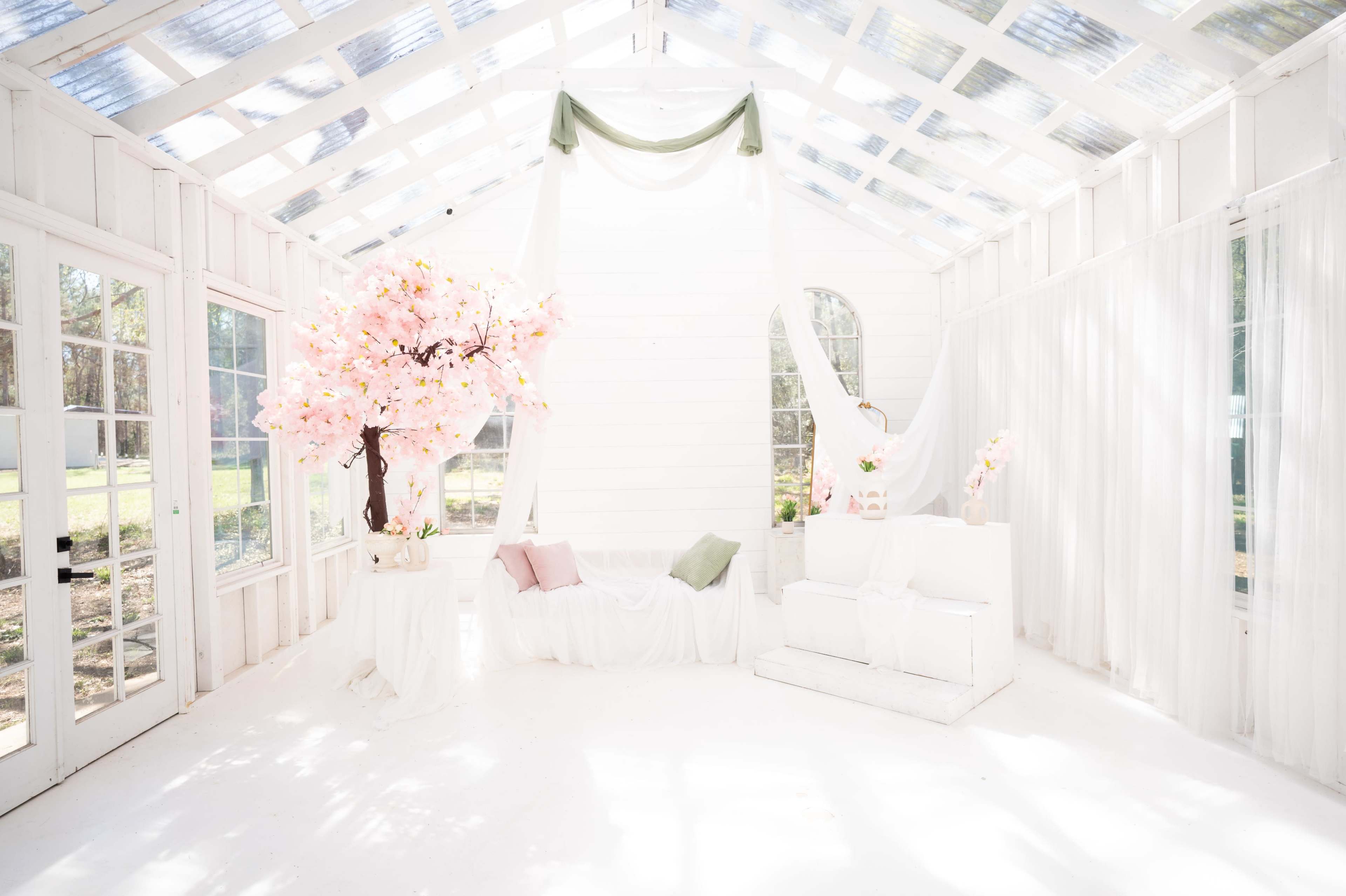 The image shows a bright, airy room with large windows, featuring a white decor, a pink cherry blossom tree, and soft furnishings.