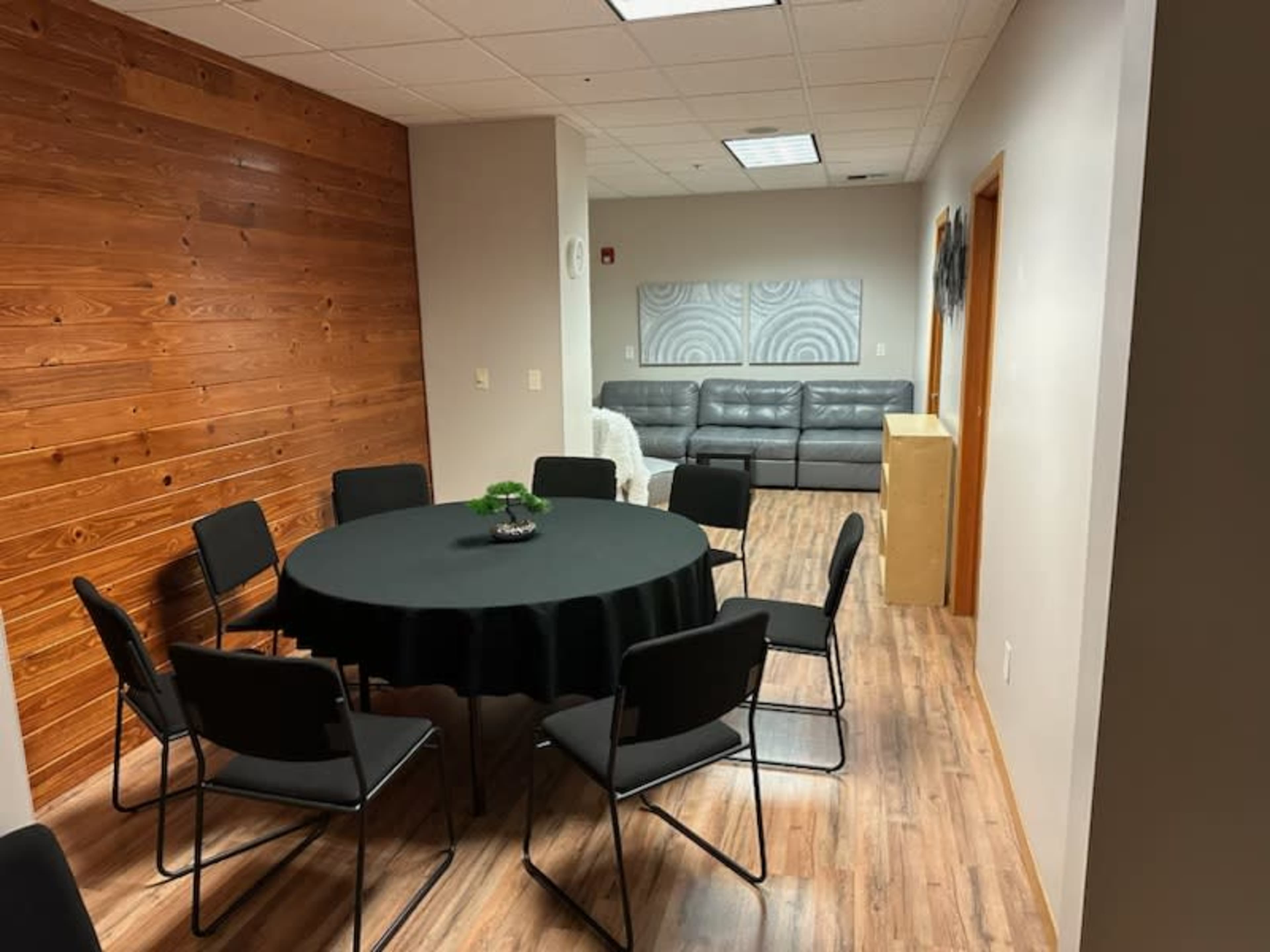 The image shows a meeting room with a round black table surrounded by chairs, a wooden accent wall, and a gray couch in the background.