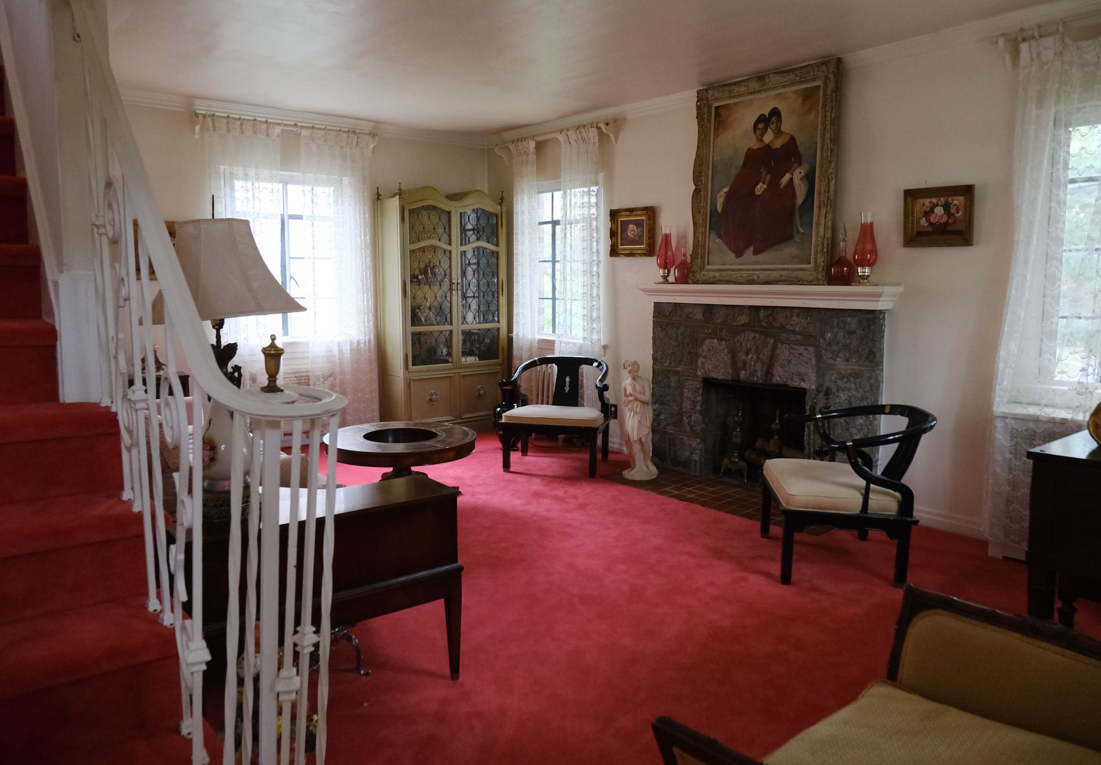 The image shows a living room featuring a red carpet, a fireplace with a stone surround, a decorative cabinet, and various pieces of furniture arranged around the space.