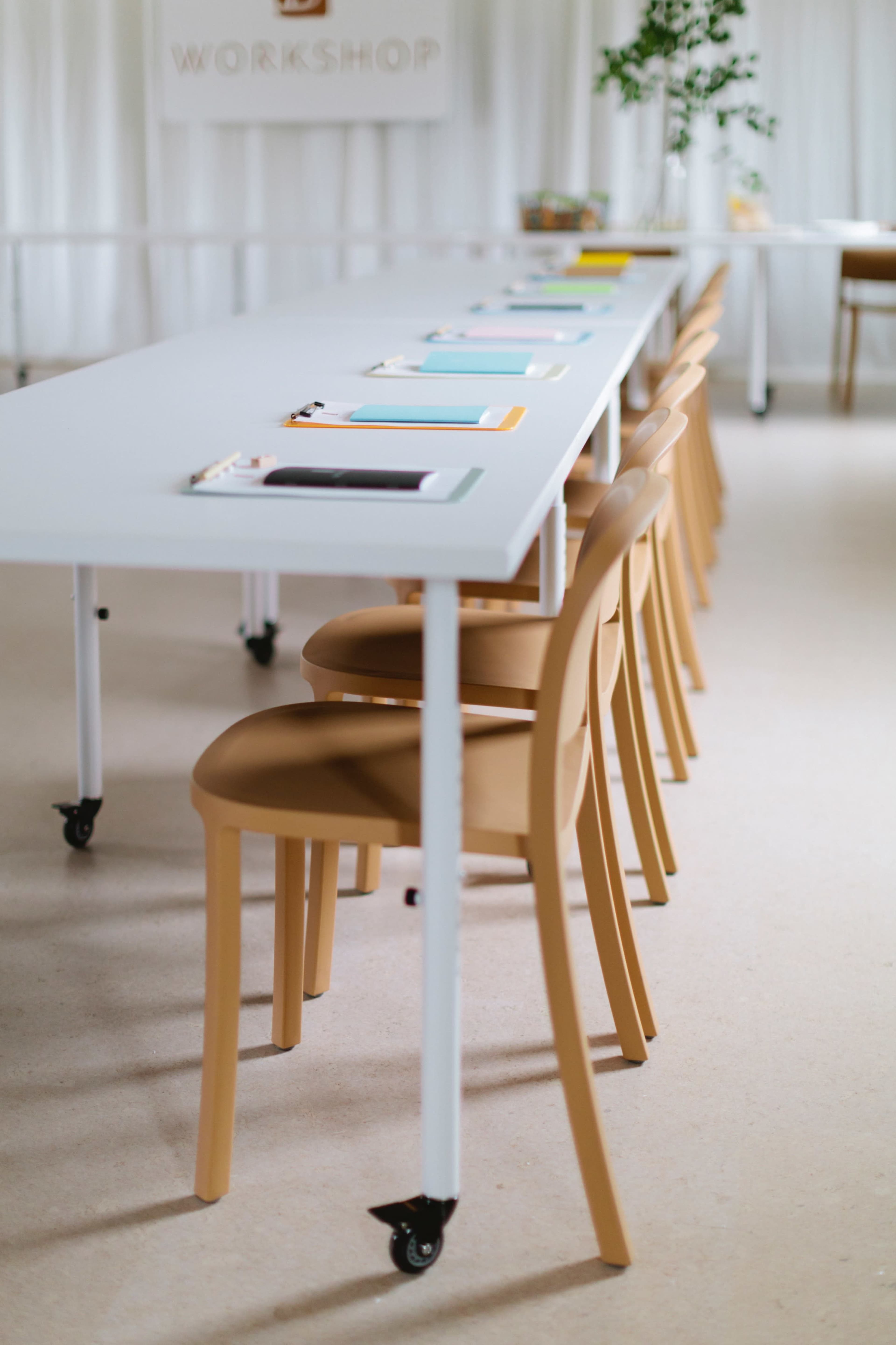 A long, minimalist table with colored notebooks and pens is arranged in a workshop space filled with light.