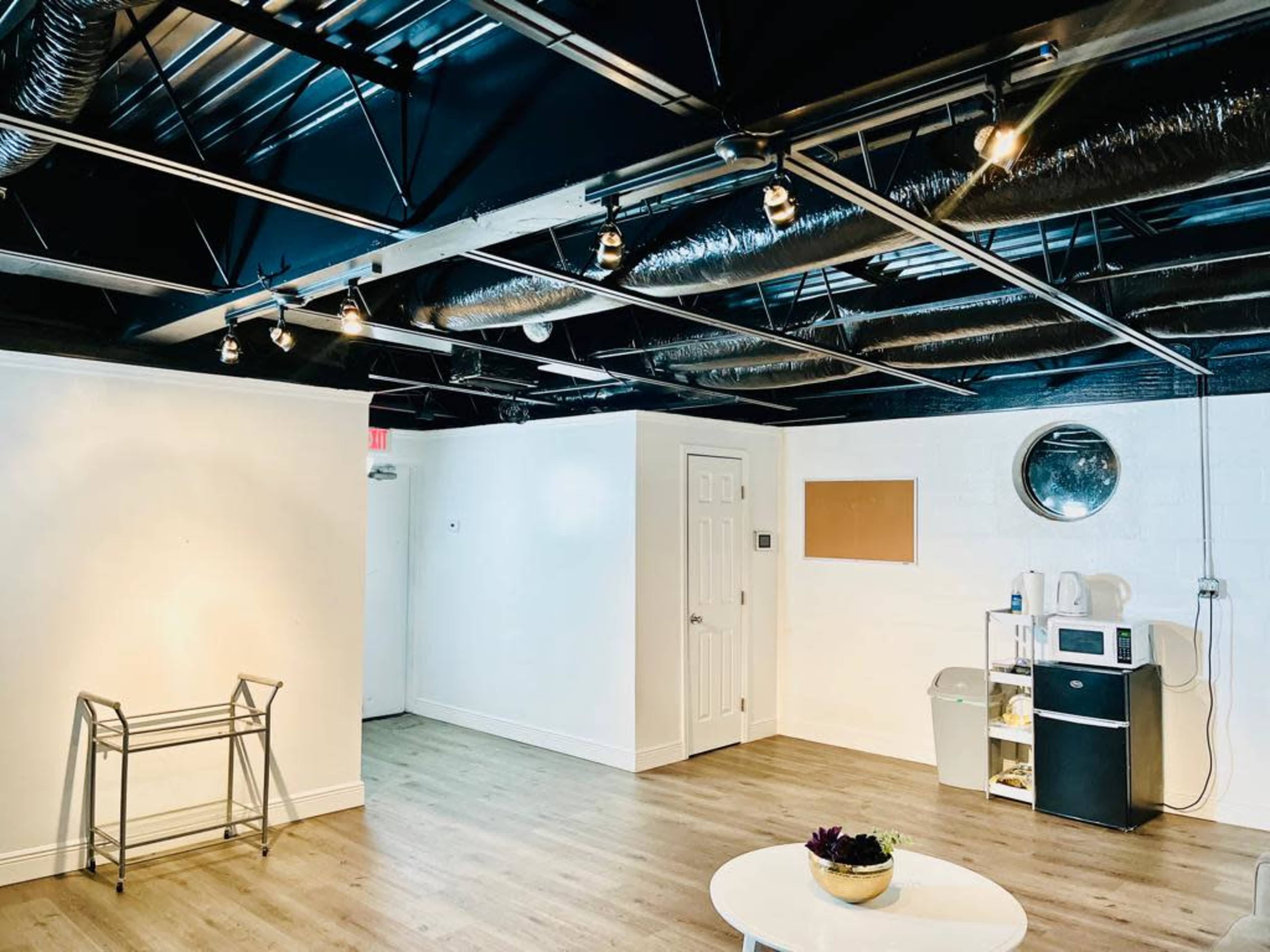 An empty room with a wooden floor, a metal frame, a door, a round mirror, a bulletin board, and kitchen appliances against one wall.