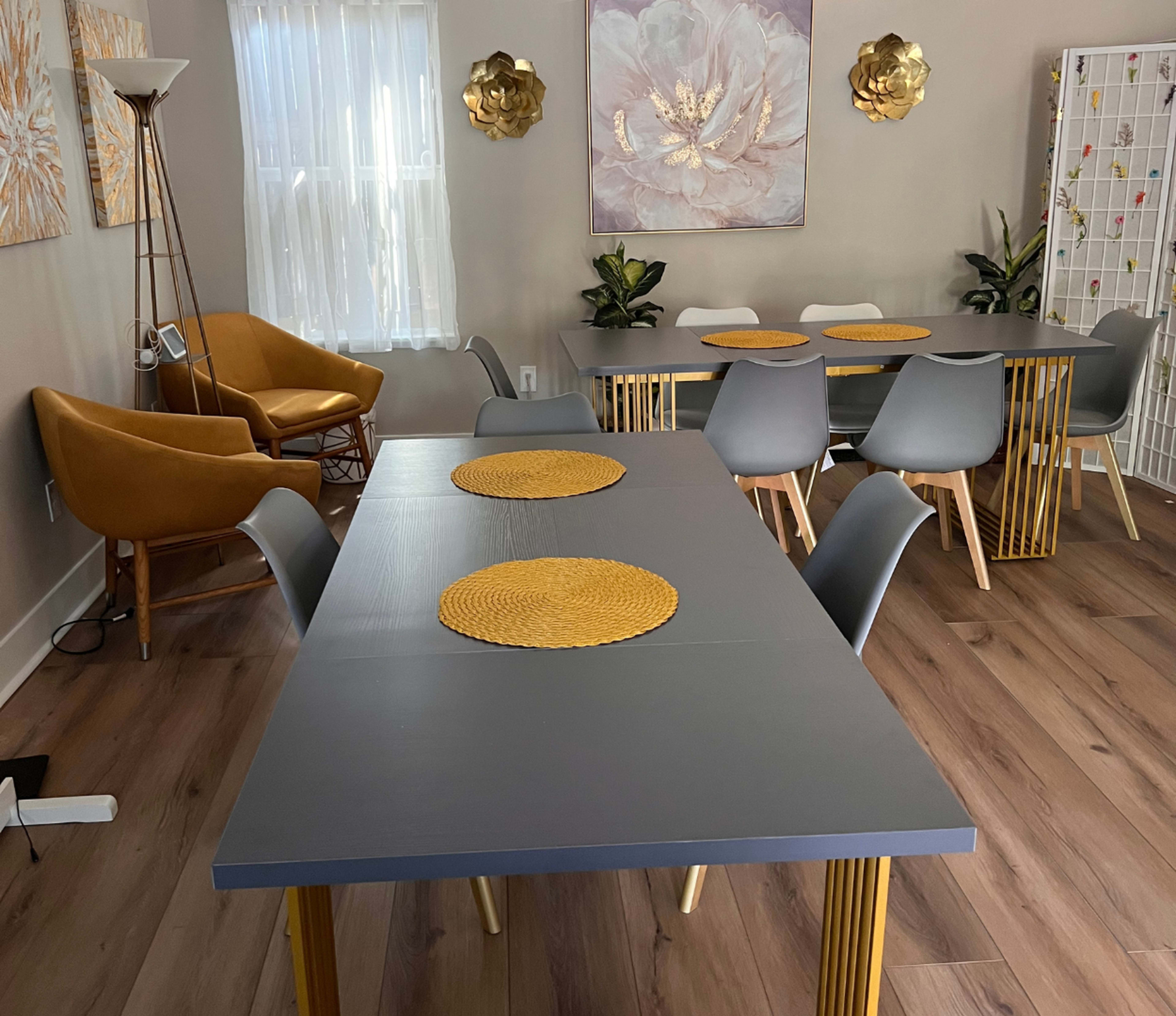 A modern dining area features a large gray table with gold legs, surrounded by several chairs, and adorned with decorative placemats, while soft light filters through a window.