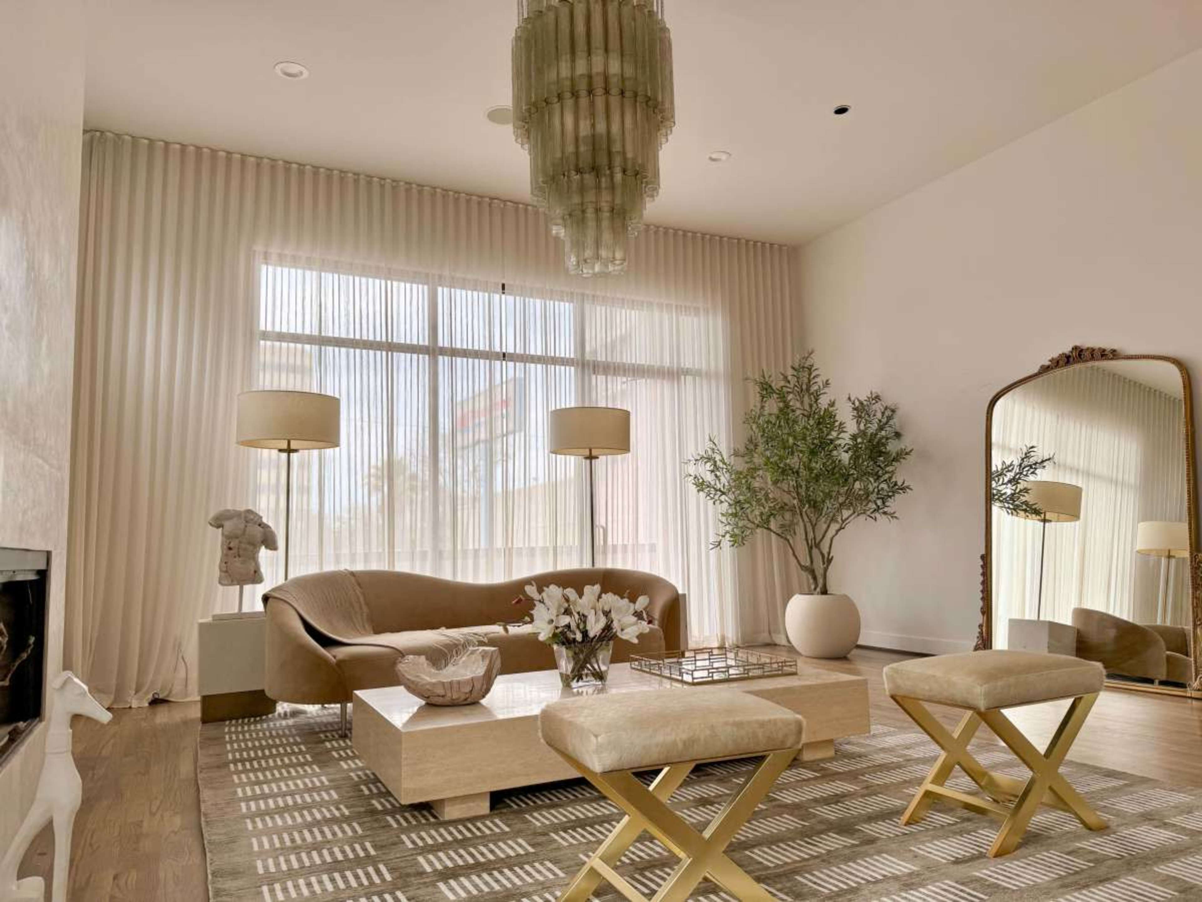 The image depicts a modern living room featuring a curved sofa, a large mirror, a coffee table with decorative items, and natural light filtered through sheer curtains.