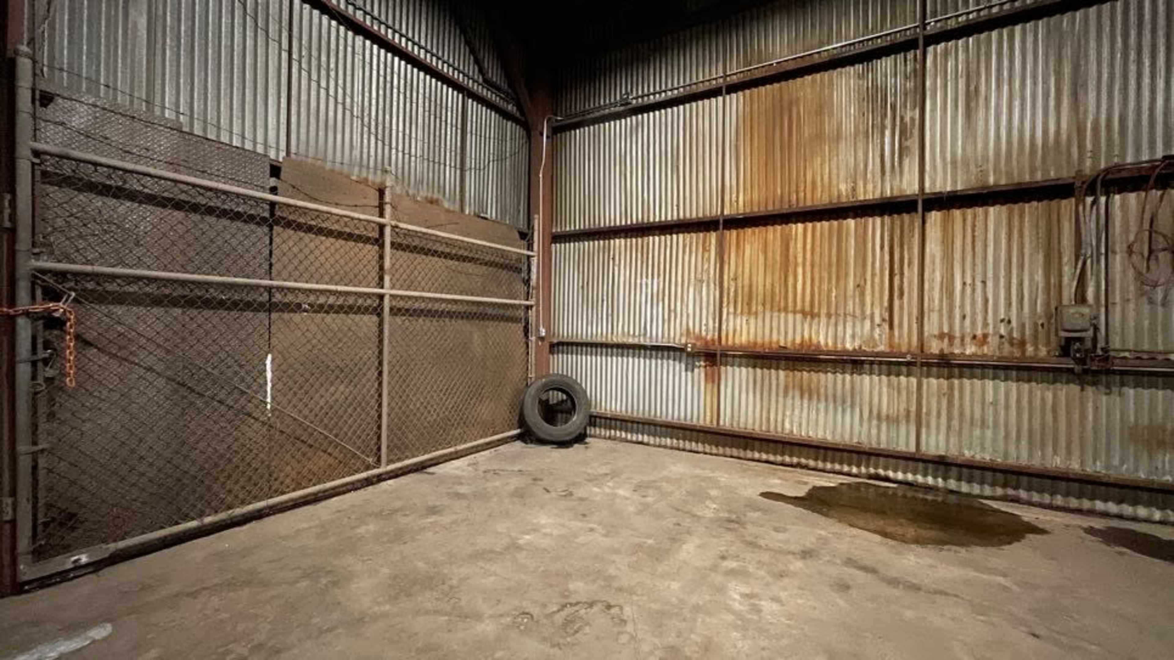 A dimly lit industrial space with corrugated metal walls, a rusted fence, and a tire in one corner.