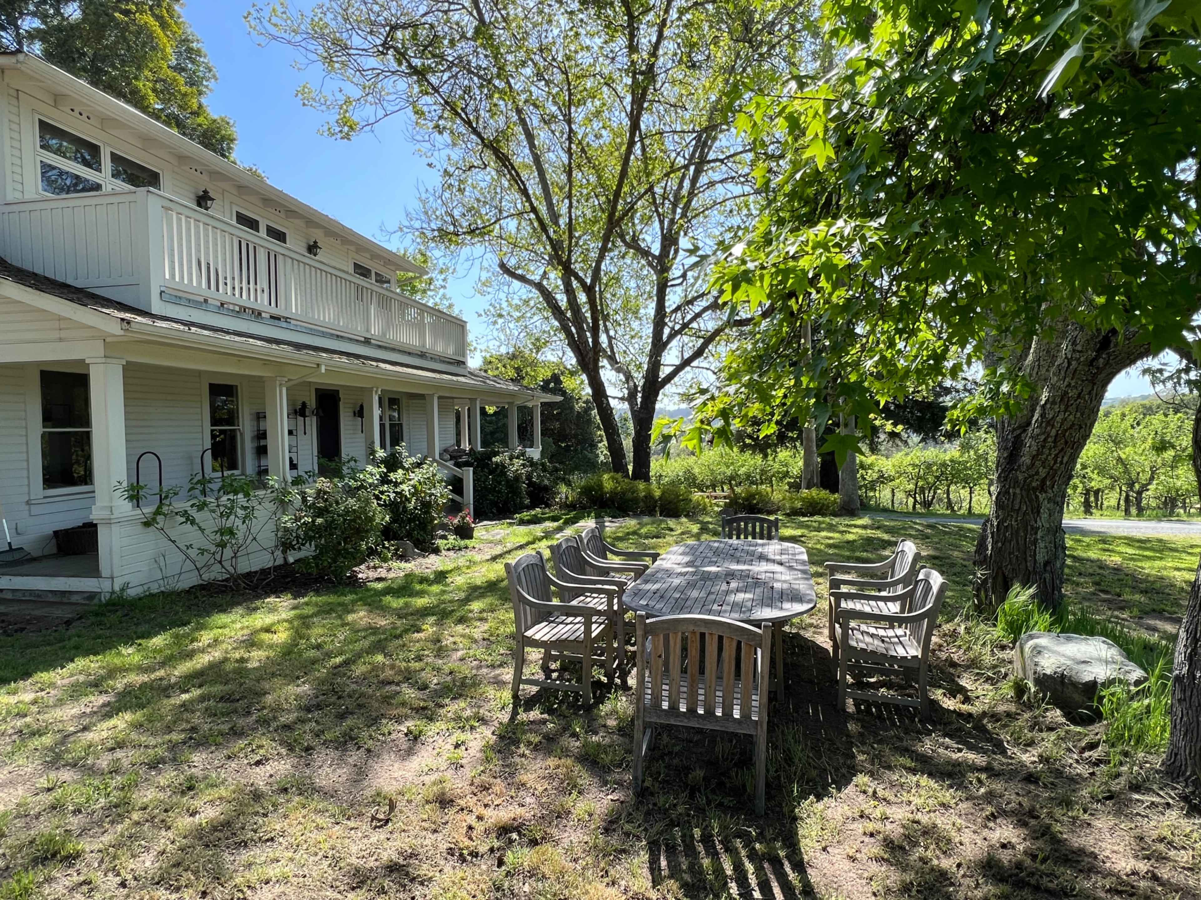A wooden outdoor dining table with eight chairs is surrounded by grass and trees near a two-story house with a balcony.