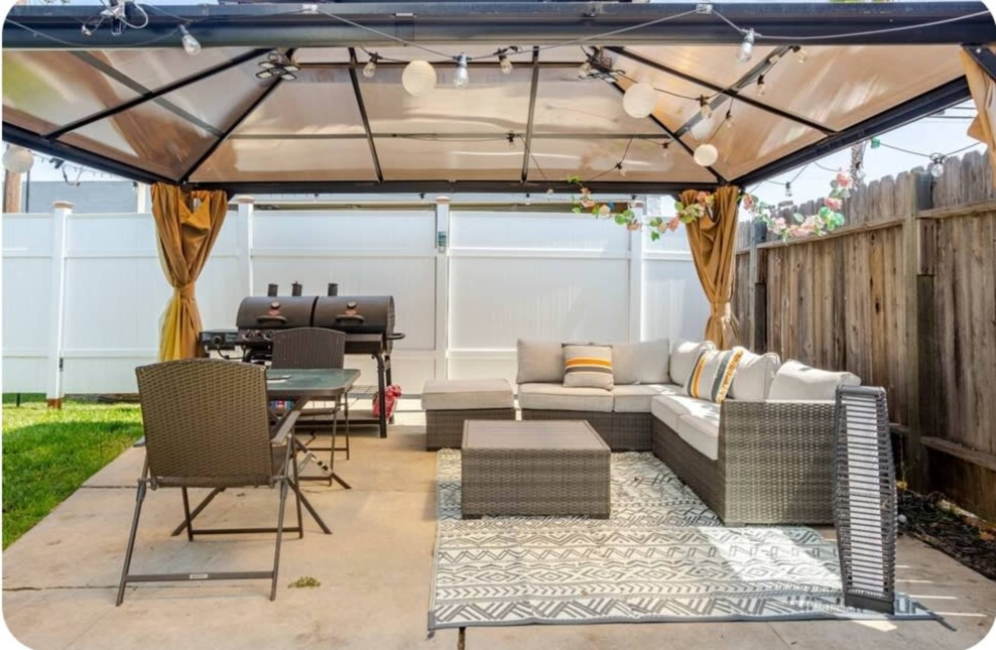 The image shows a covered outdoor seating area with a sofa set, a dining table, and a barbecue grill under a gazebo.
