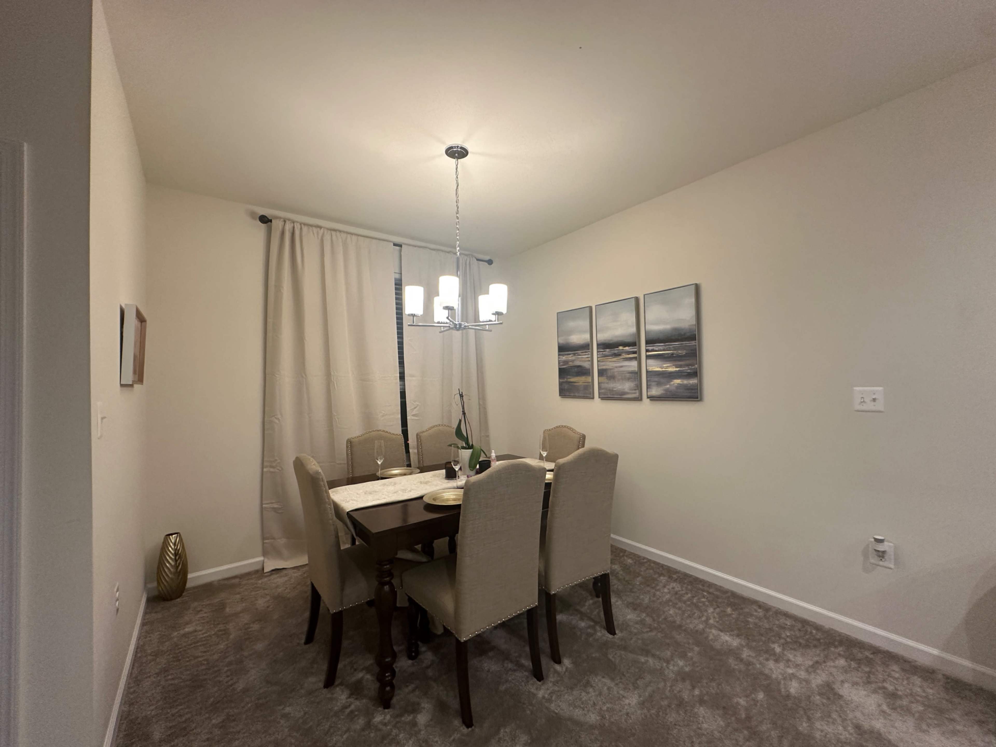 A dining room features a wooden table surrounded by six upholstered chairs, with a chandelier above and three landscape paintings on the wall.