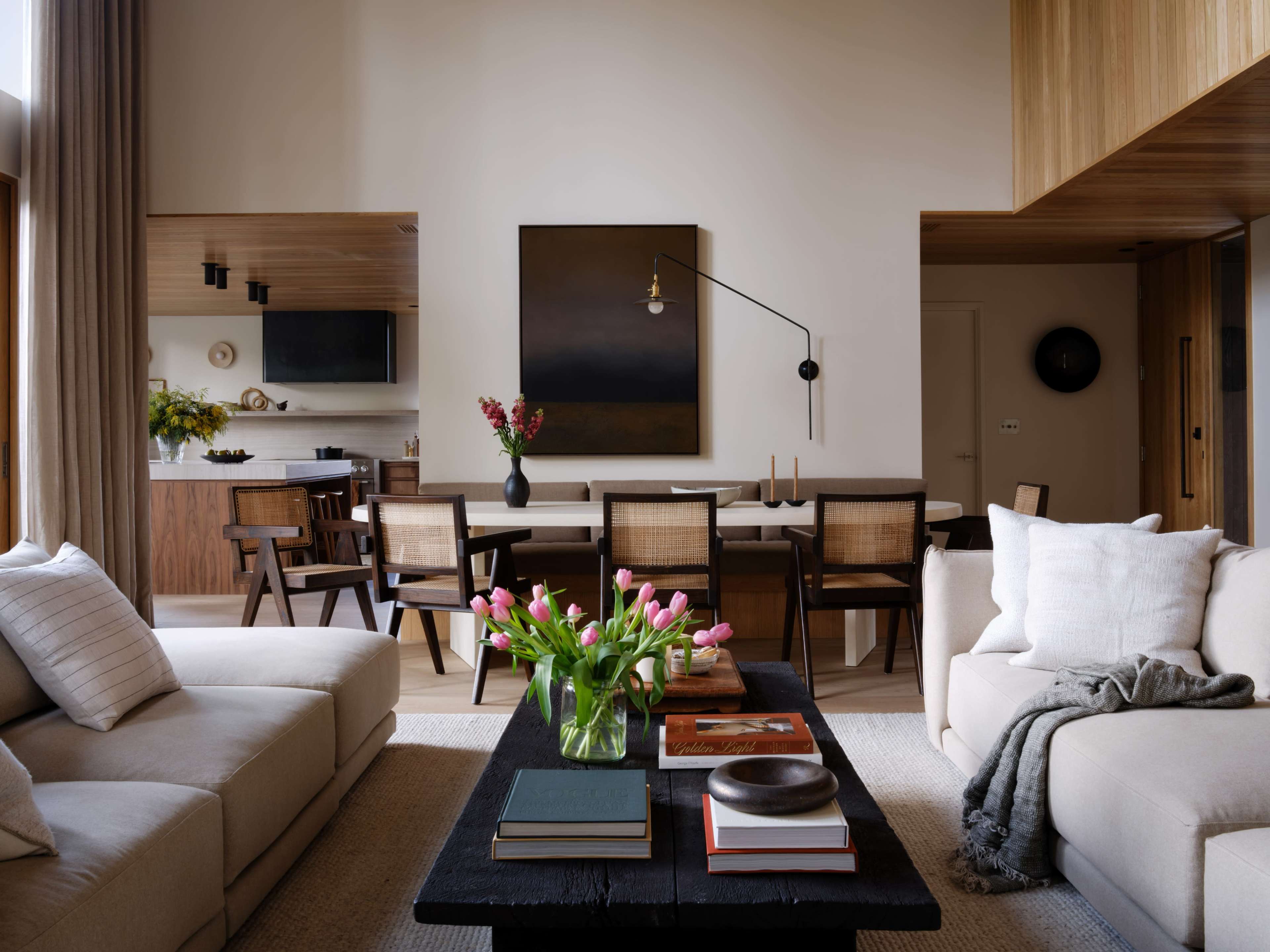 The image shows a modern living room with a beige sofa, a coffee table filled with books, and a dining area featuring a wooden table and chairs.