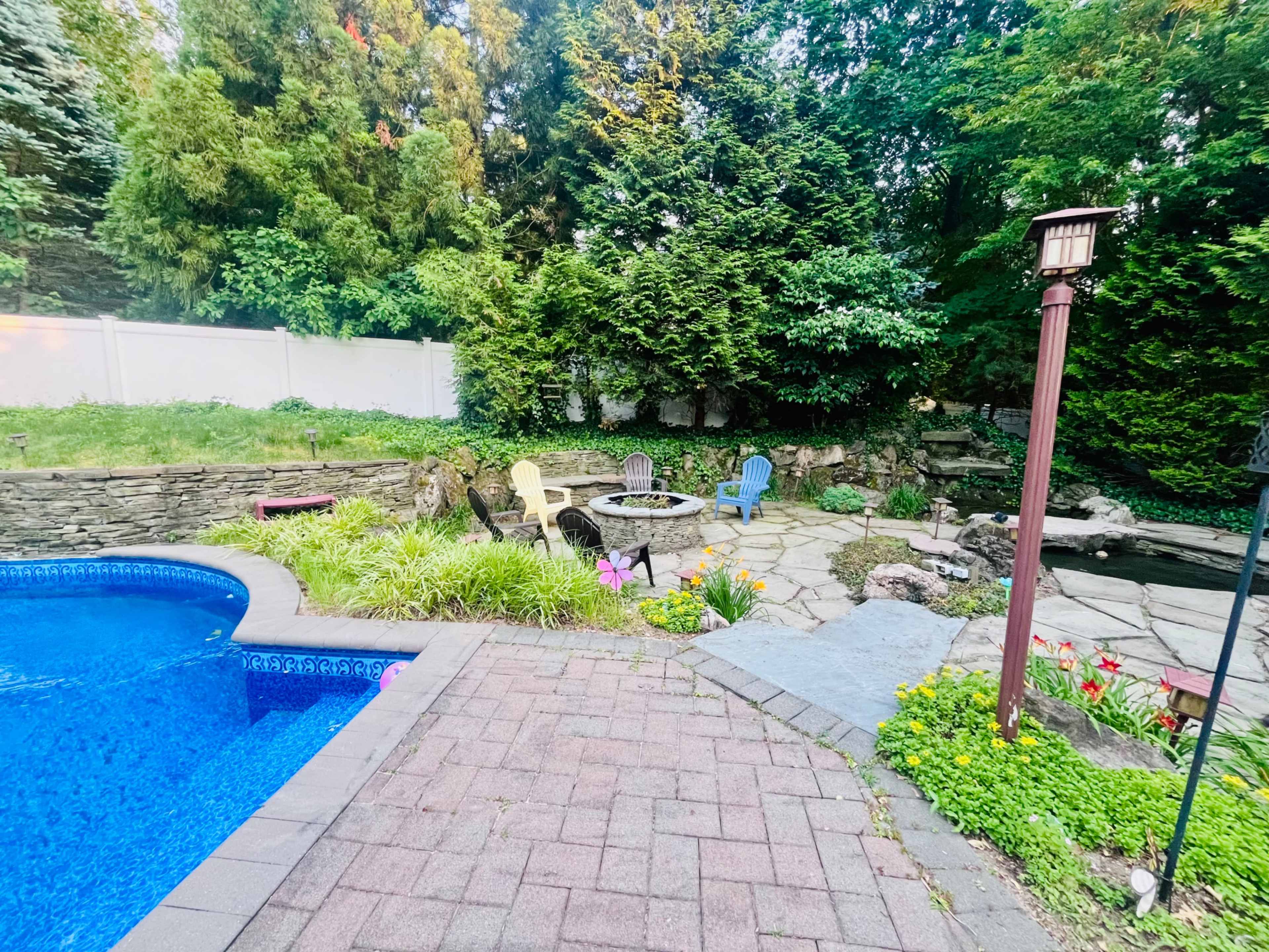 A stone patio area featuring a firepit surrounded by colorful chairs and lush greenery, adjacent to a swimming pool.