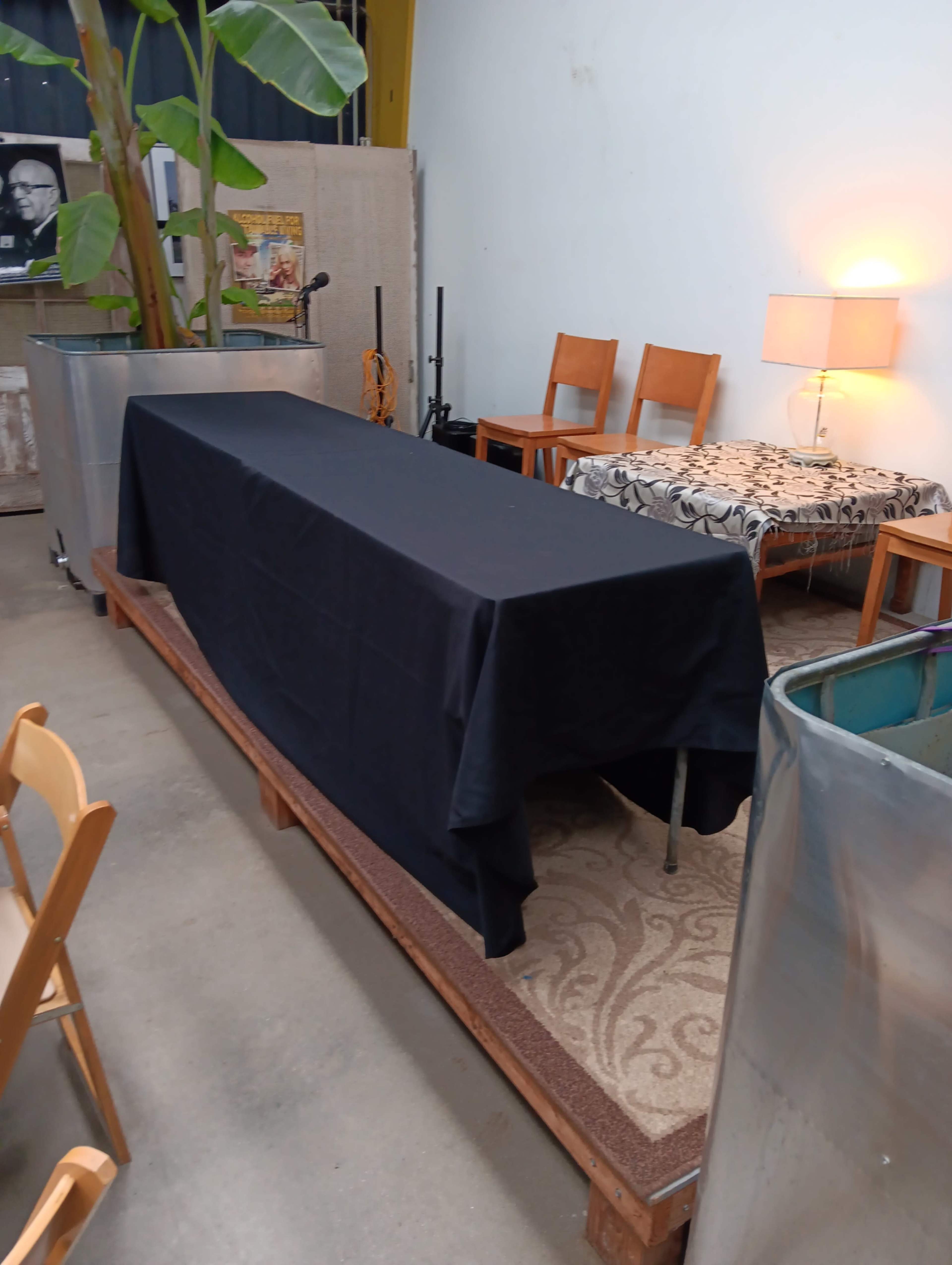 A room with a long table covered by a black cloth, surrounded by wooden chairs, a lamp, and two metal containers.